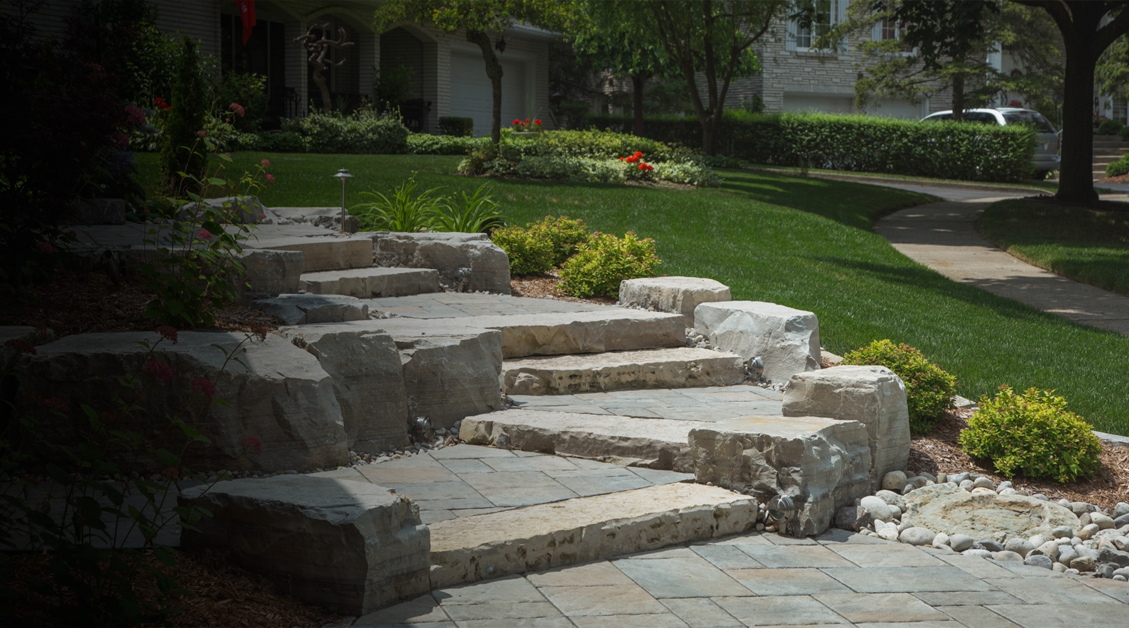 The image shows a landscaped garden with a stone stairway, amid lush plants, leading to a well-manicured lawn in front of residential homes.