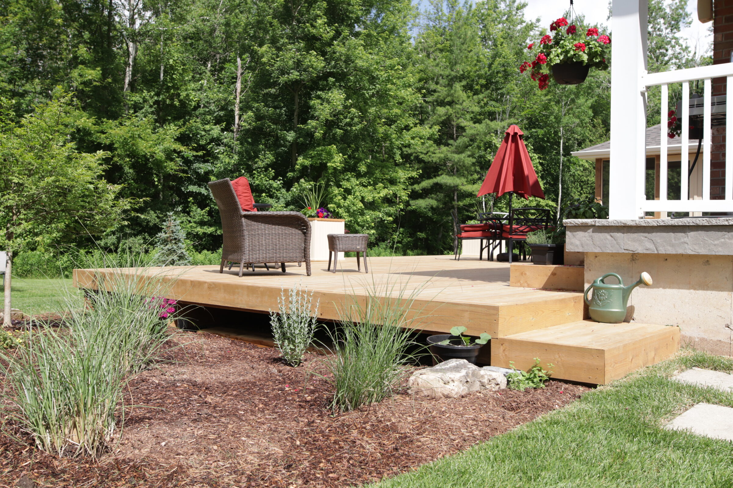 A spacious wooden deck with outdoor furniture and a red umbrella, surrounded by greenery, mulch, and a water pitcher decoration.