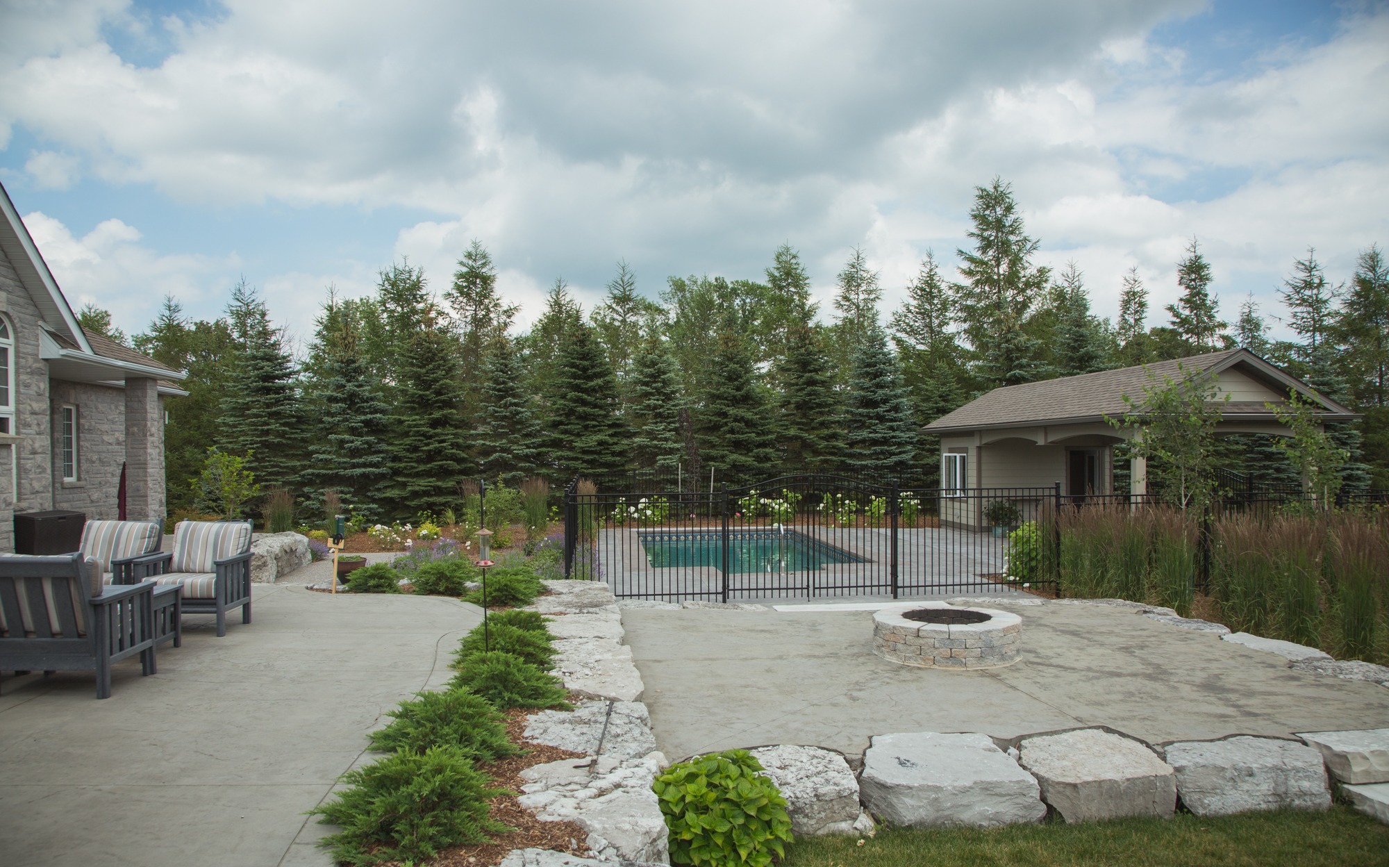 A well-maintained backyard with stone paving, outdoor furniture, a fire pit, tall trees, and a view of a fenced pool beside a small house.