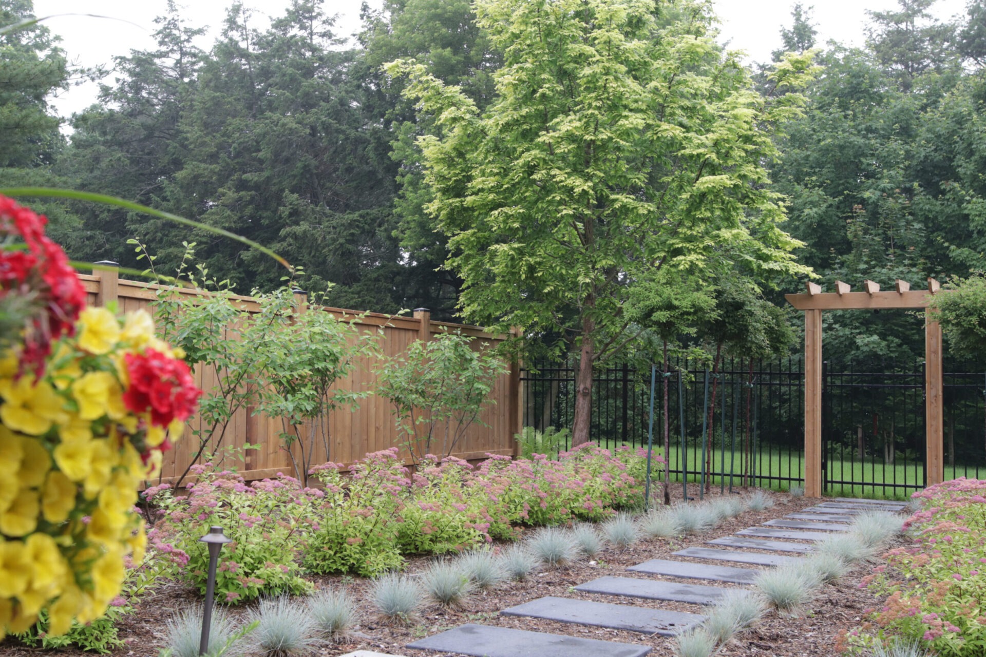 A serene garden pathway with large stepping stones, surrounded by flowering shrubs, a wooden fence, and a lush treeline in the background.