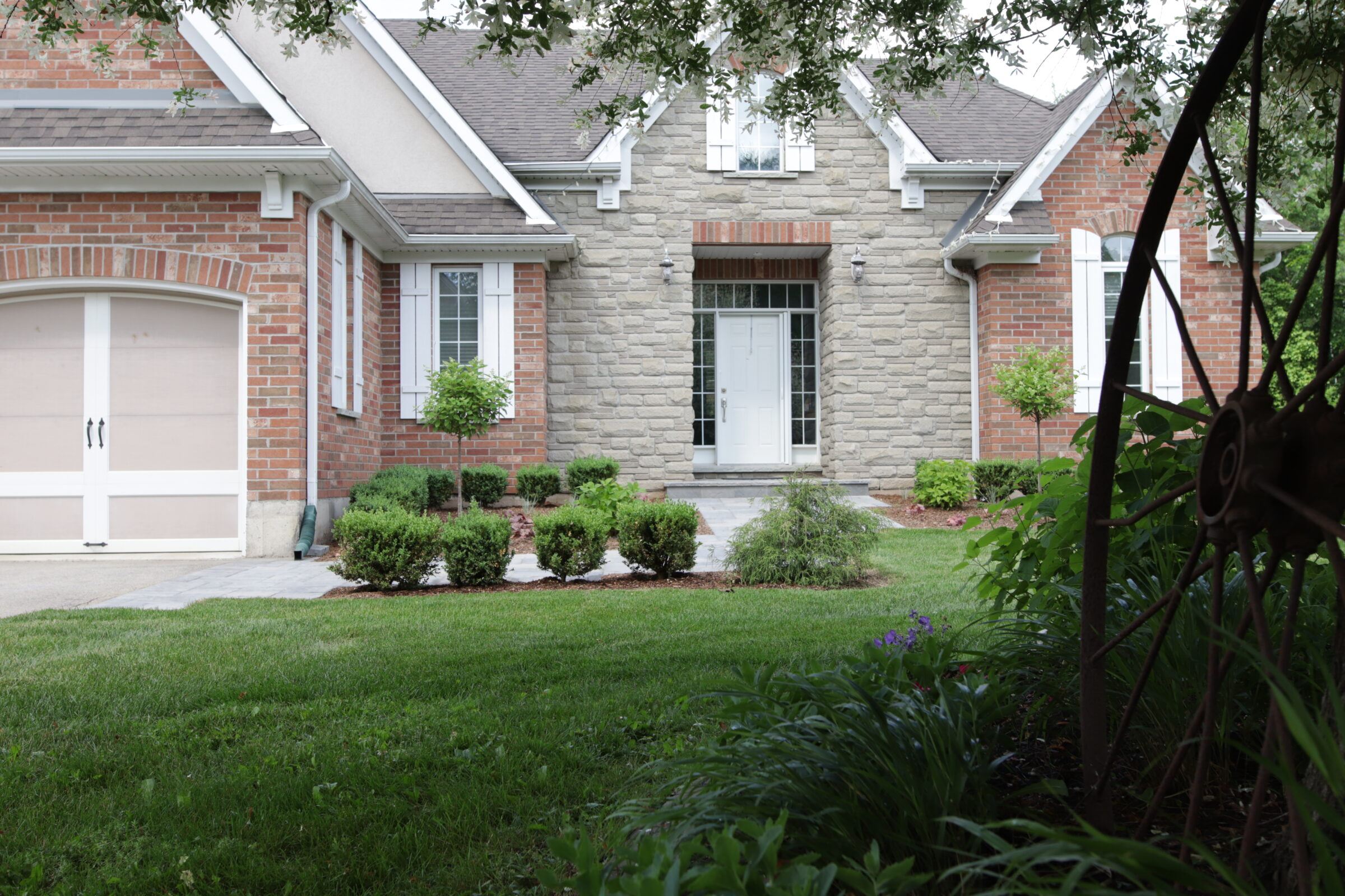 This is a suburban brick house with a stone facade, white garage door, a manicured lawn, and neatly trimmed bushes. There's a garden in the foreground.