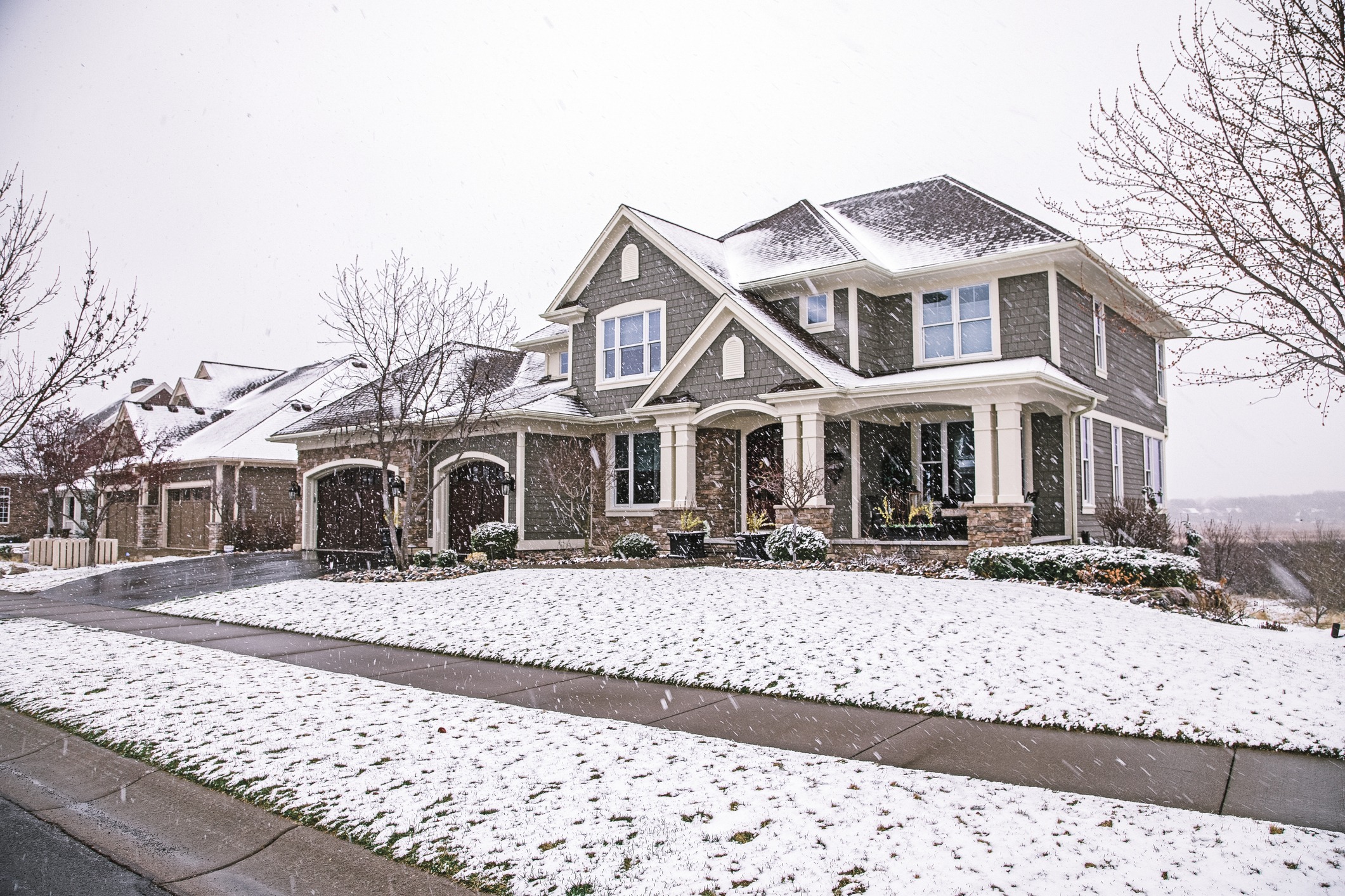 A two-story suburban house with a brown and gray exterior under a light snowfall, featuring a covered porch, snow-dusted lawn, and bare trees.