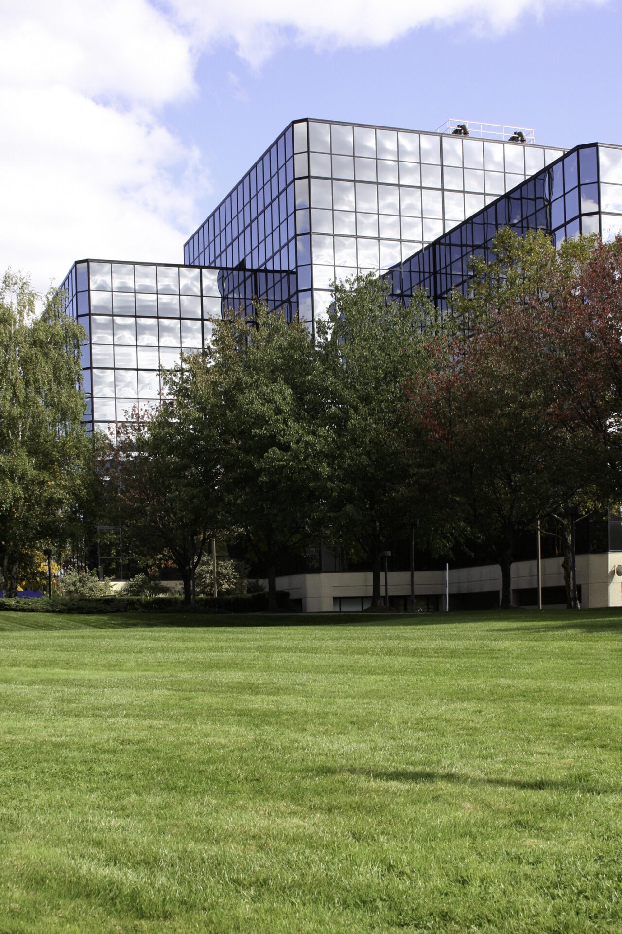 A modern glass office building overlooks a manicured lawn with trees showing early autumn colors under a partly cloudy sky.