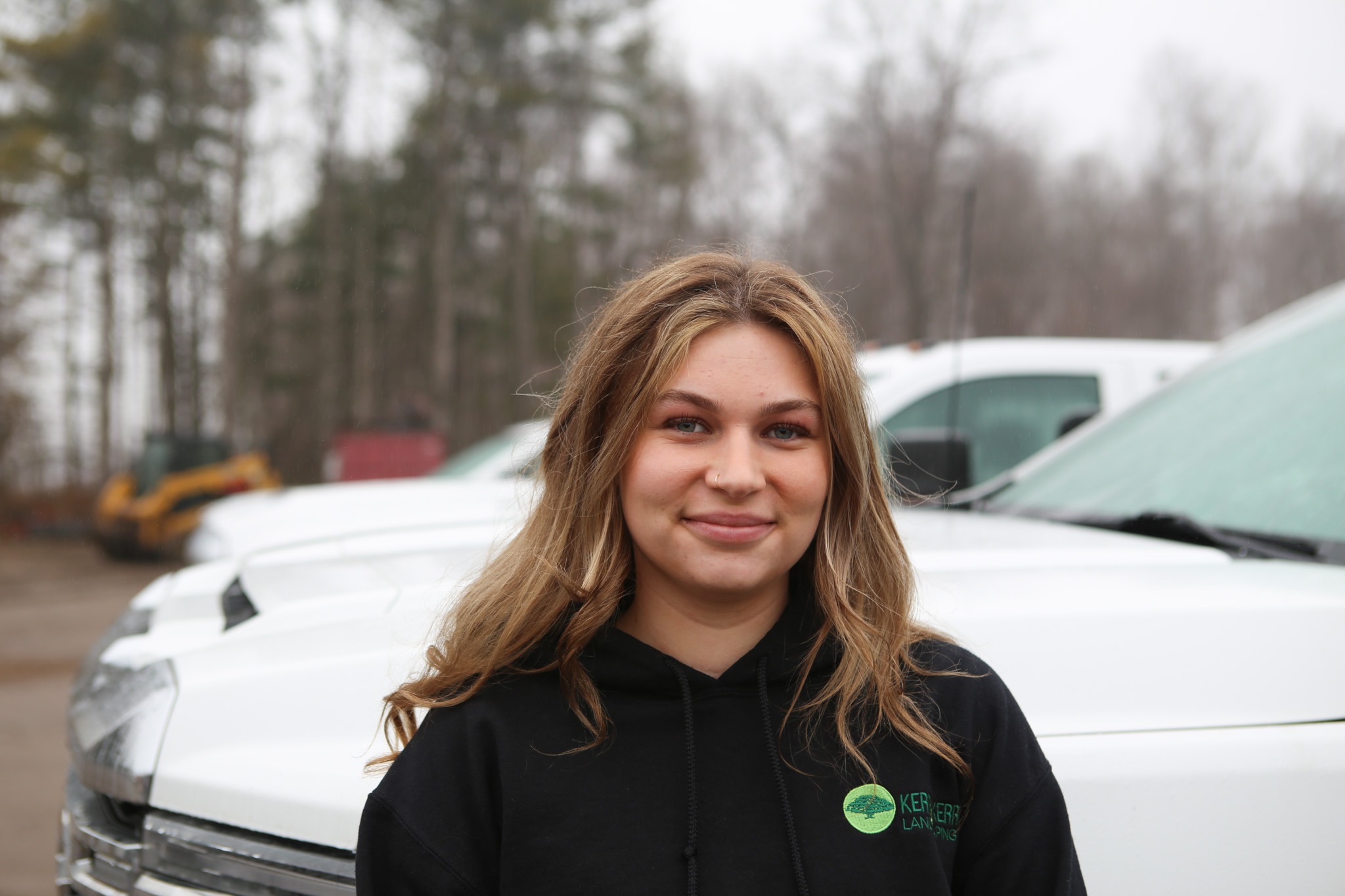 A person with blonde hair smiling at the camera, wearing a black hoodie, with white vans and trees in the blurred background.