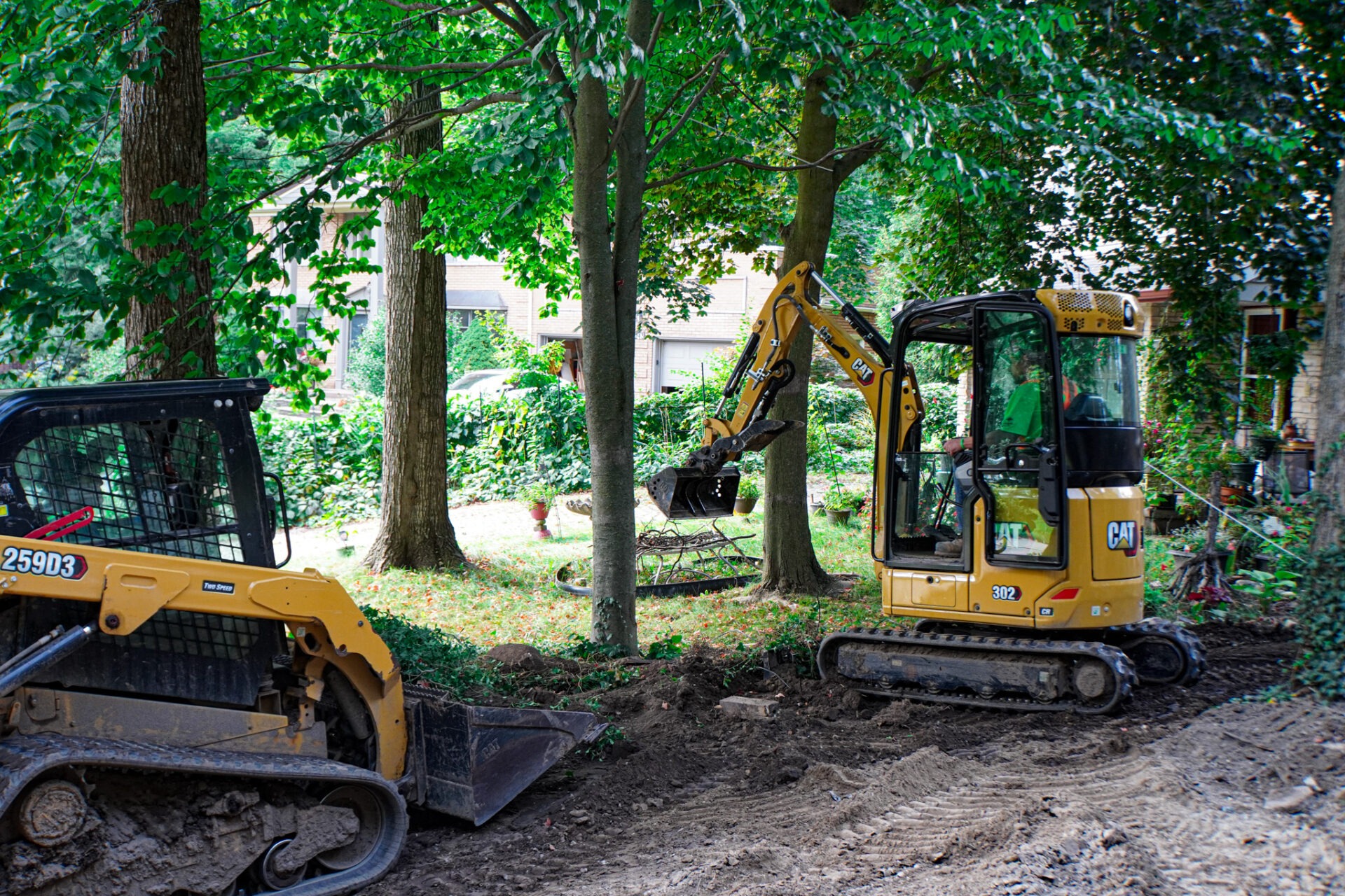 A yellow mini excavator and a skid steer loader are working in a wooded area with freshly moved earth and green trees surrounding the construction site.