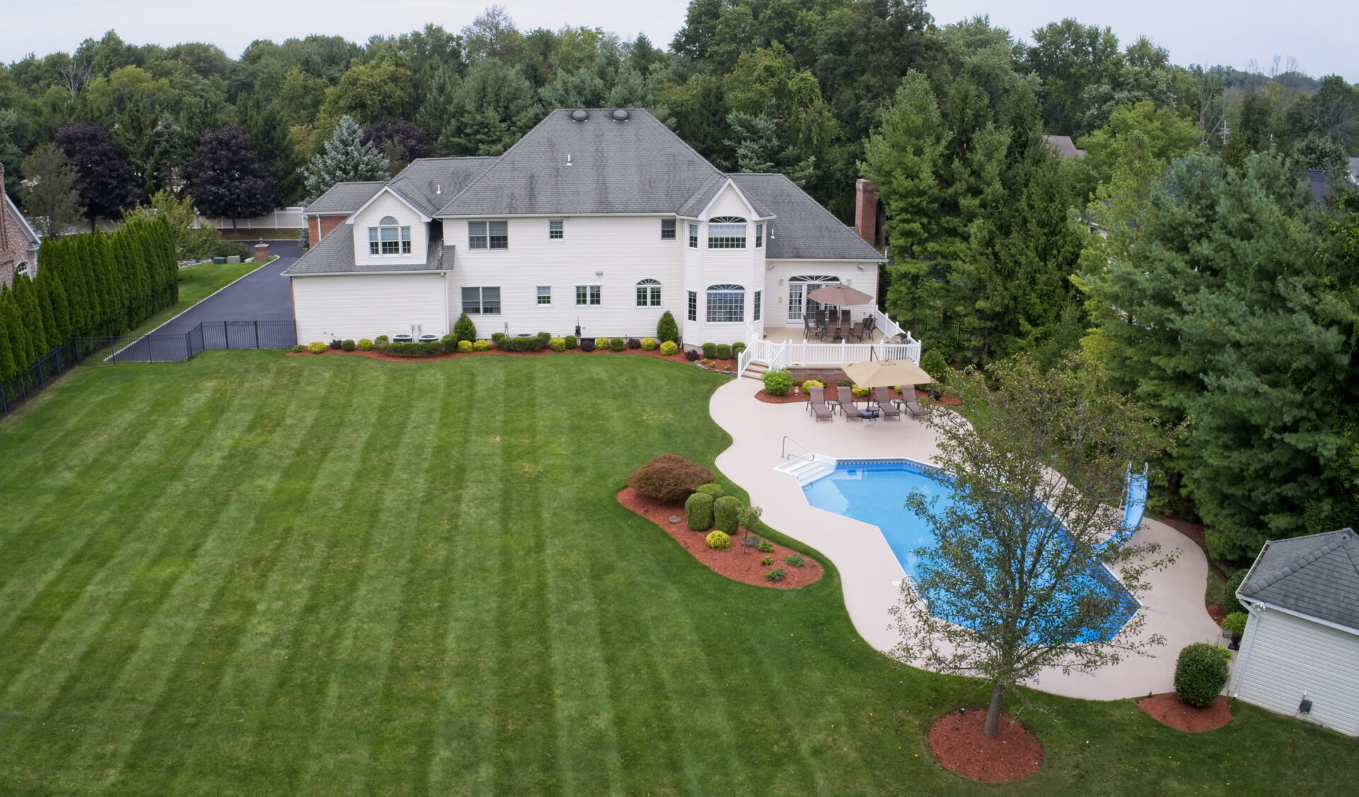 This is an aerial view of a large two-story house with a well-manicured lawn, an in-ground swimming pool, and surrounded by trees.