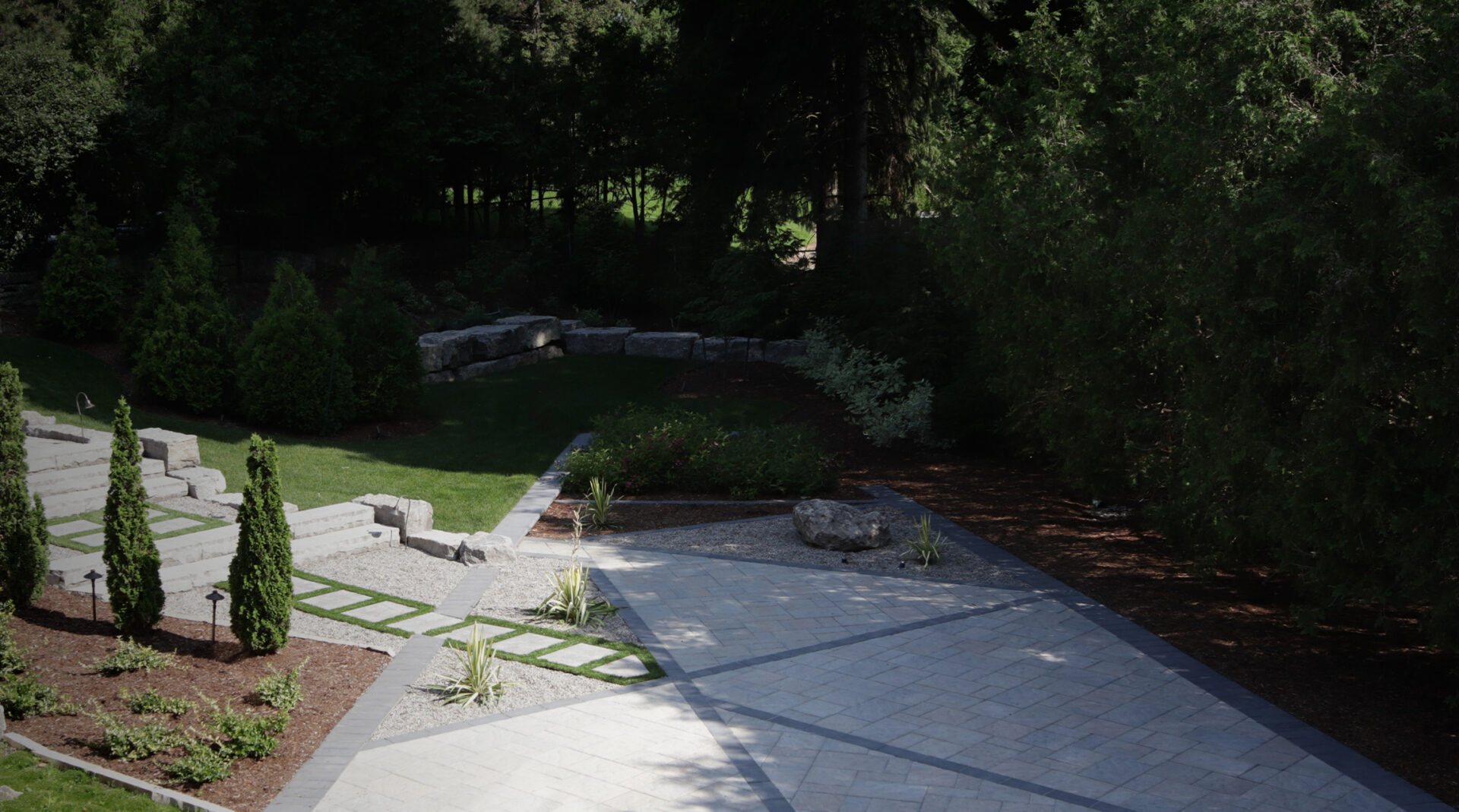 This image shows a landscaped garden with a stone pathway, manicured bushes, columns of conical trees, and a surrounding backdrop of dense trees.