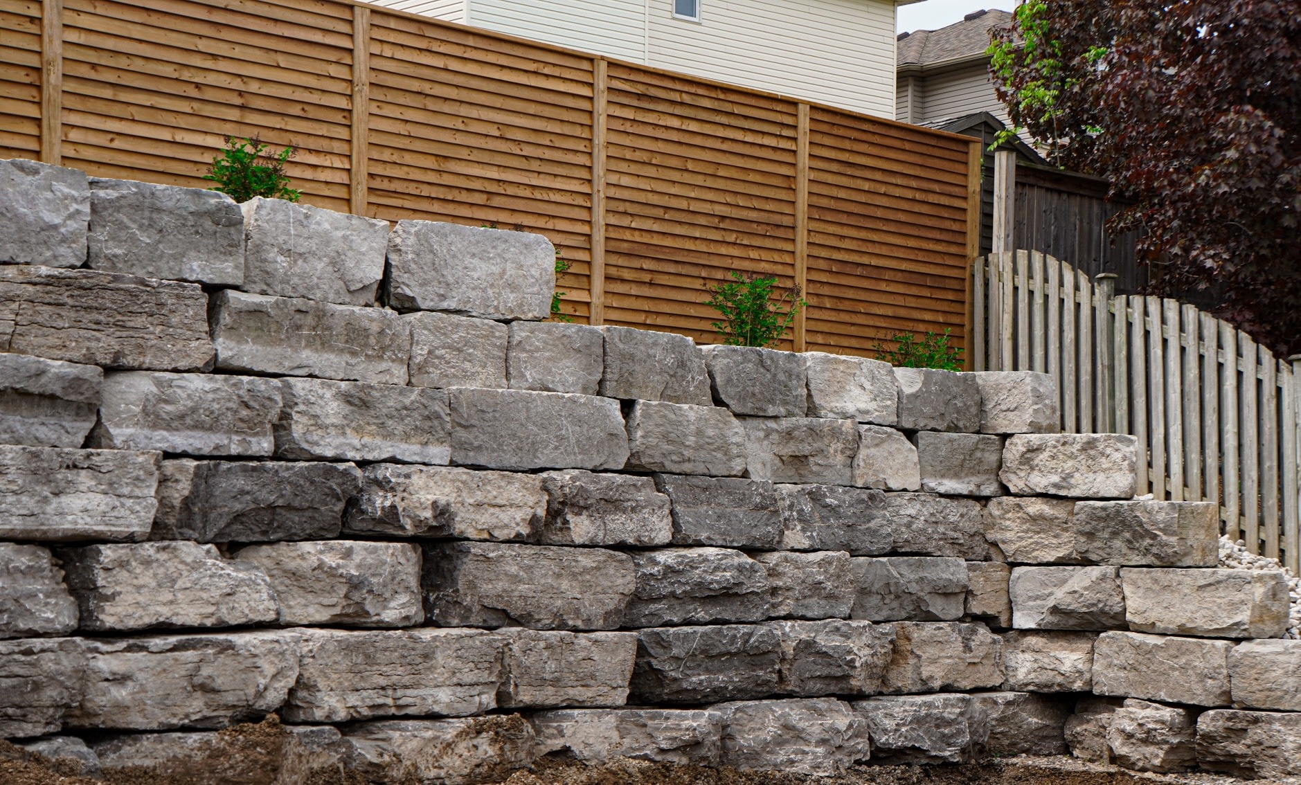 A stacked stone retaining wall in a residential area with a wooden fence and some green shrubs visible behind the wall.