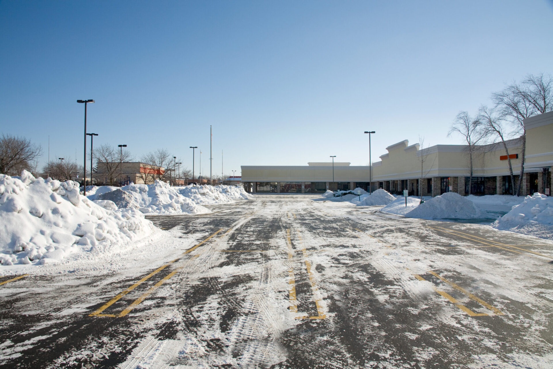 A snowy parking lot with large piles of plowed snow, bare trees, a strip mall in the background, and clear blue skies.