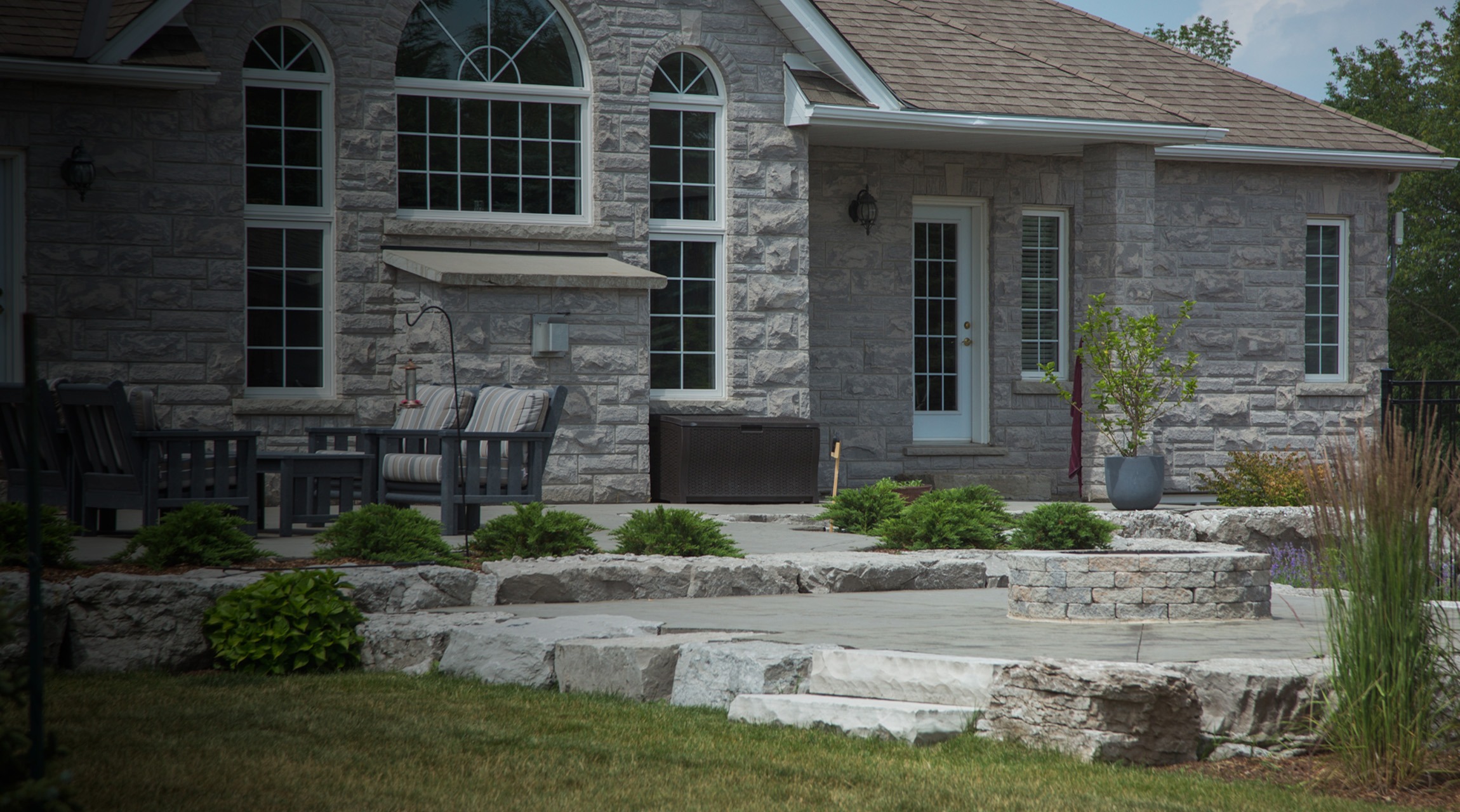 This image shows a stone house with large windows, a patio with outdoor furniture, and layers of landscaped stone steps leading to a lawn.