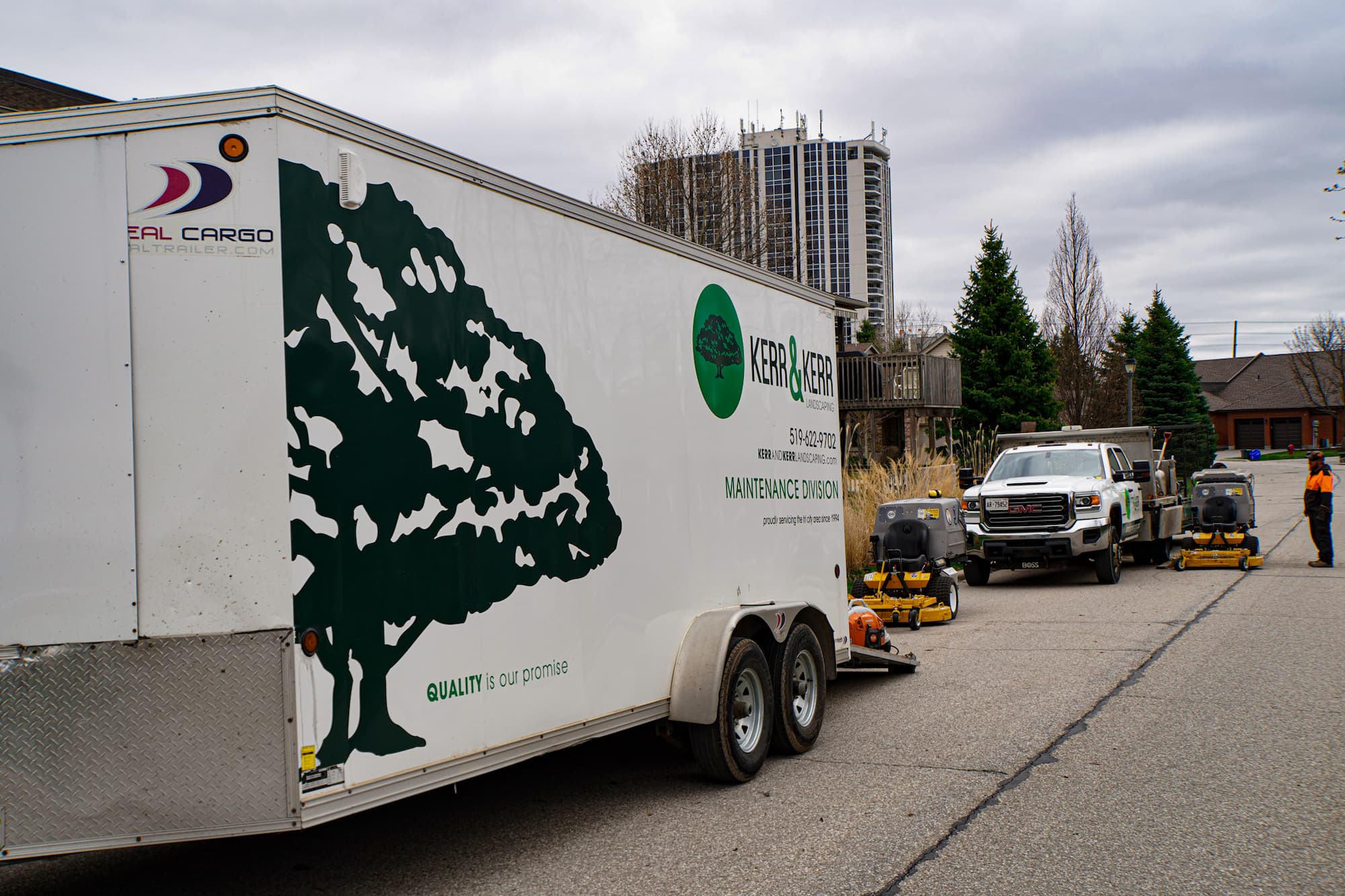 A landscape trailer with a tree logo is parked on a street. A person in reflective clothing works near mowing equipment and a white pickup truck.