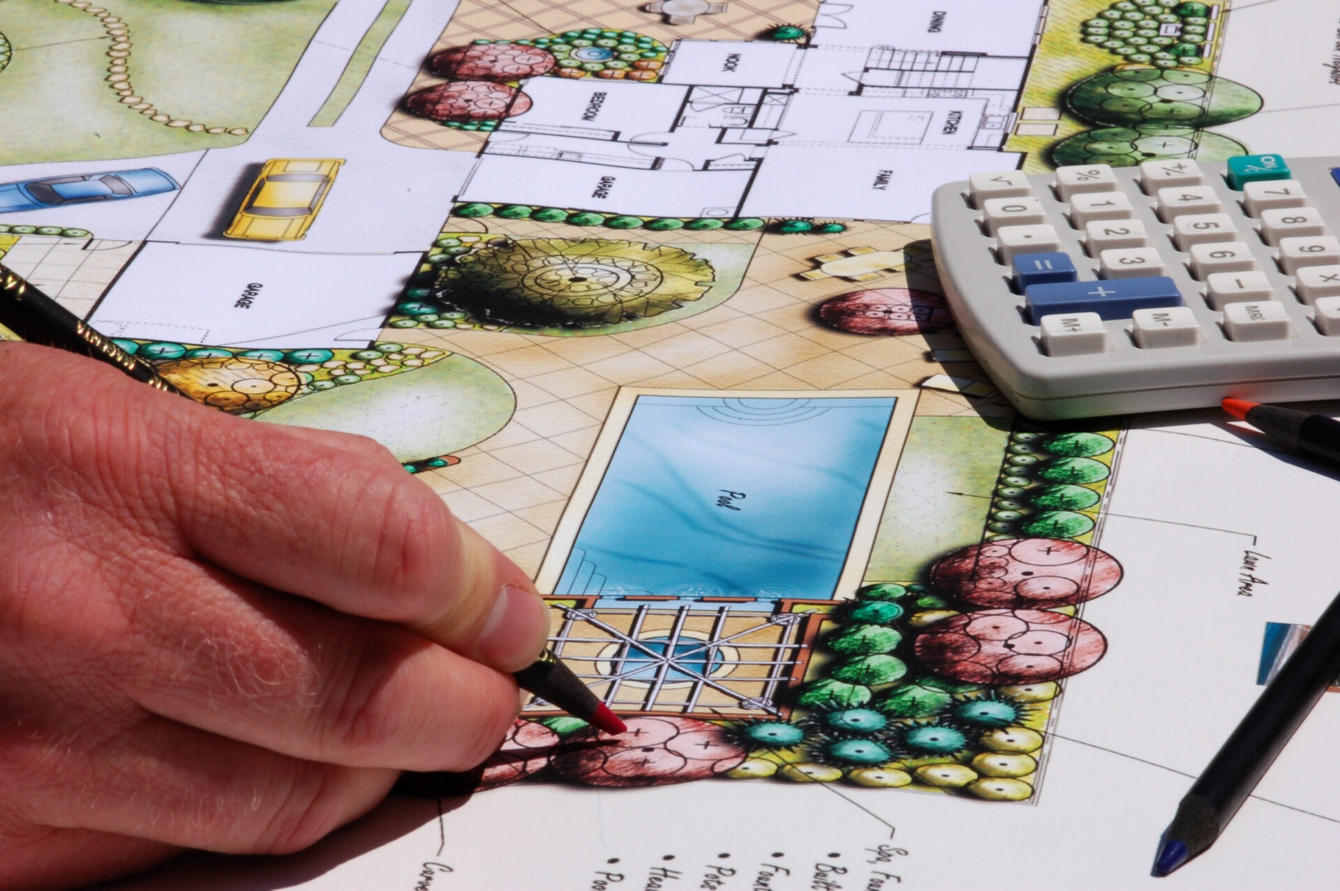 A person's hand holds a pencil over a colorful landscape architecture plan, featuring a pool, trees, and garden areas, alongside a calculator.