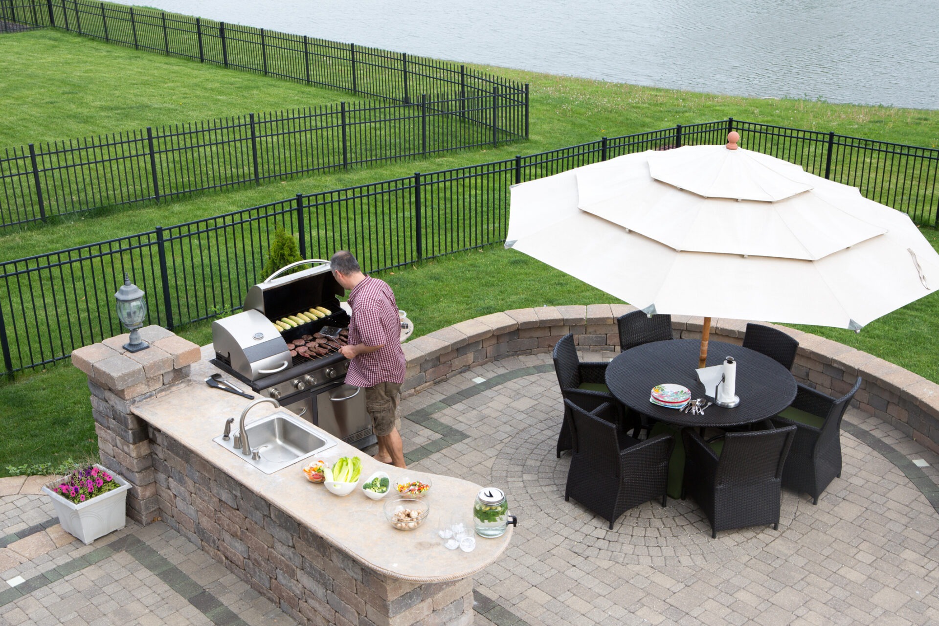 A person is grilling food outdoors next to a body of water, with a patio dining set under an umbrella, and a well-maintained lawn behind a fence.