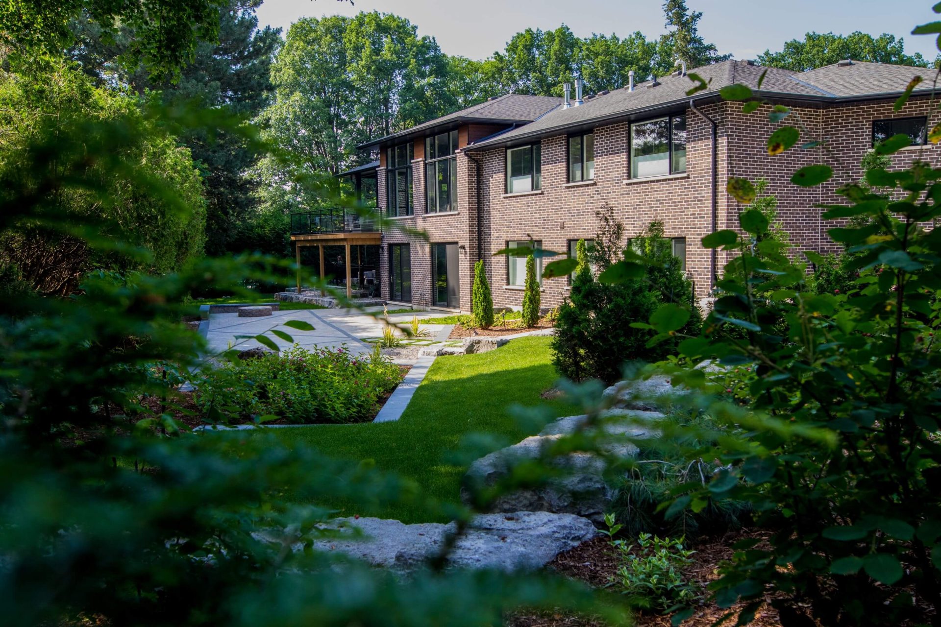 A modern two-story brick house with large windows and a balcony, surrounded by a lush garden, green lawn, and a stone walkway in a serene setting.
