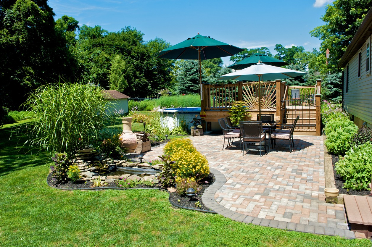 A well-manicured garden with a paver patio, chairs, umbrellas, and a deck. A water feature, lush plants, and a green lawn enhance the tranquil outdoor space.
