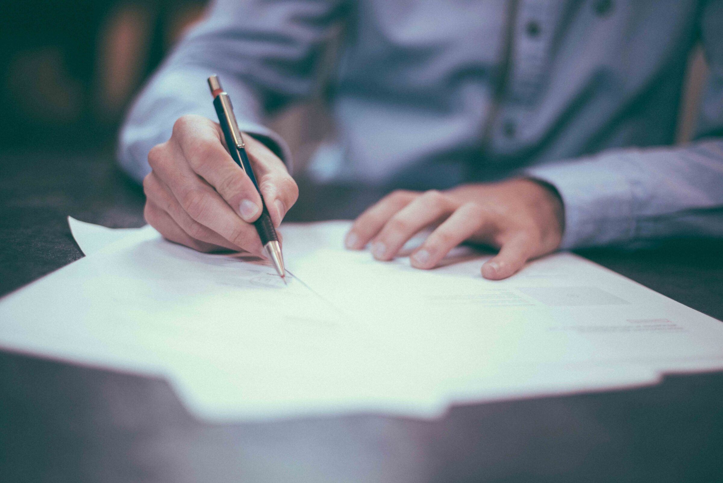 The image shows a person writing on paper with a pen, focusing on the hands and the writing task, with a blurred background enhancing concentration.