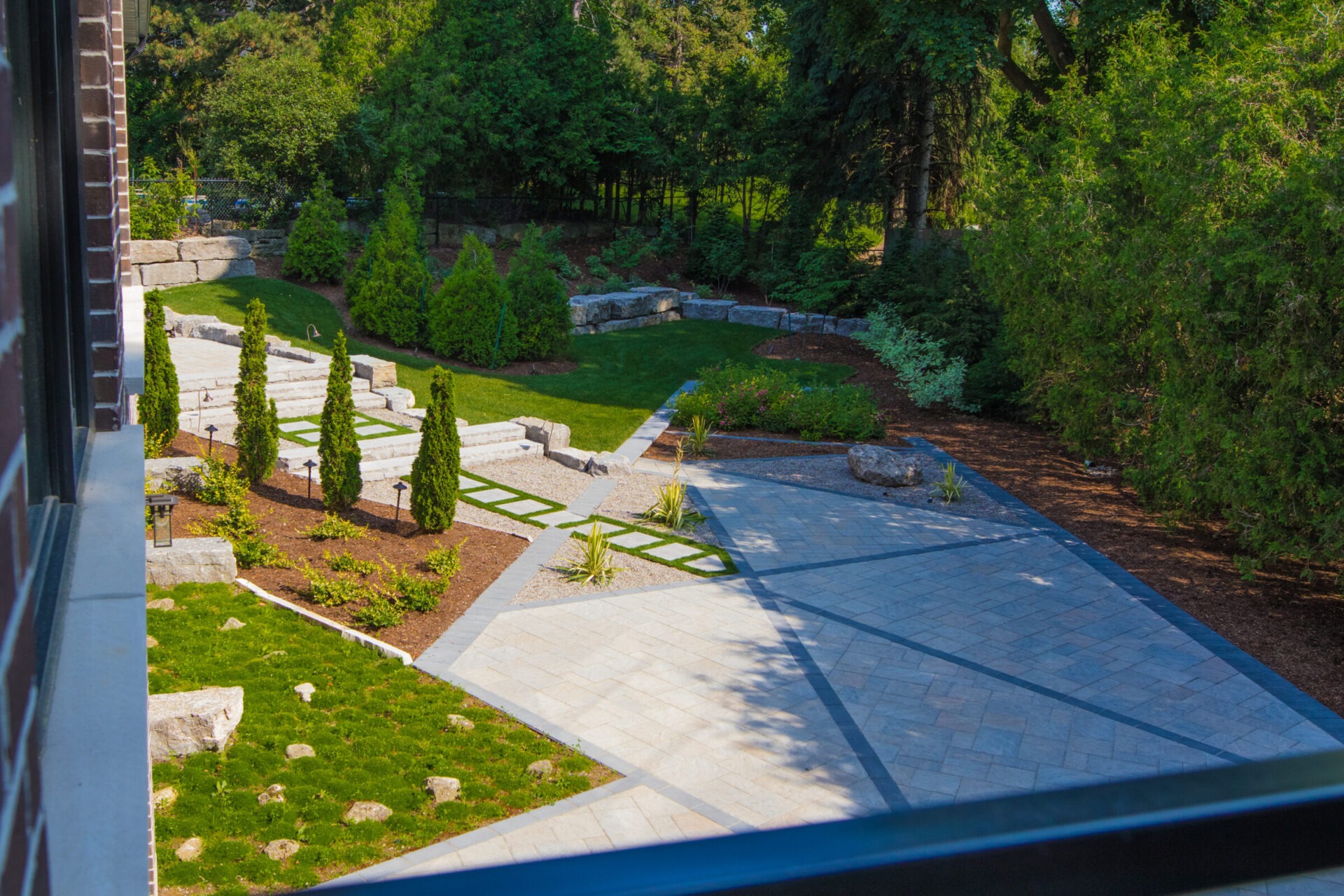 Modern backyard landscape featuring stone pavers, geometric lawn inlays, garden beds, and mature trees surrounding the property.