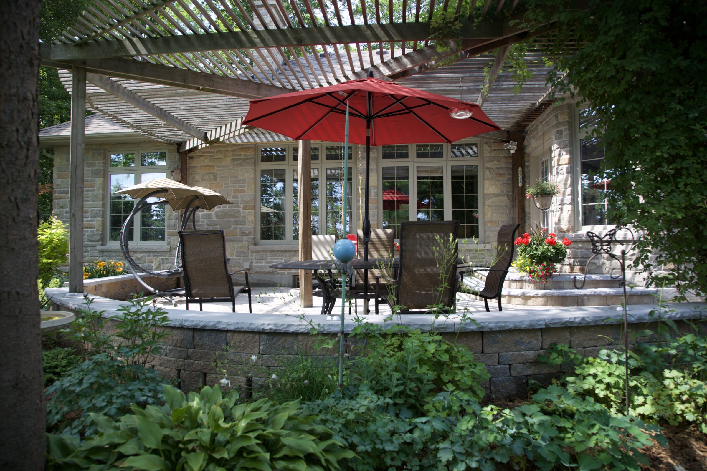 Backyard stone patio with red umbrella, outdoor dining set, and pergola, surrounded by lush greenery and flowering plants.
