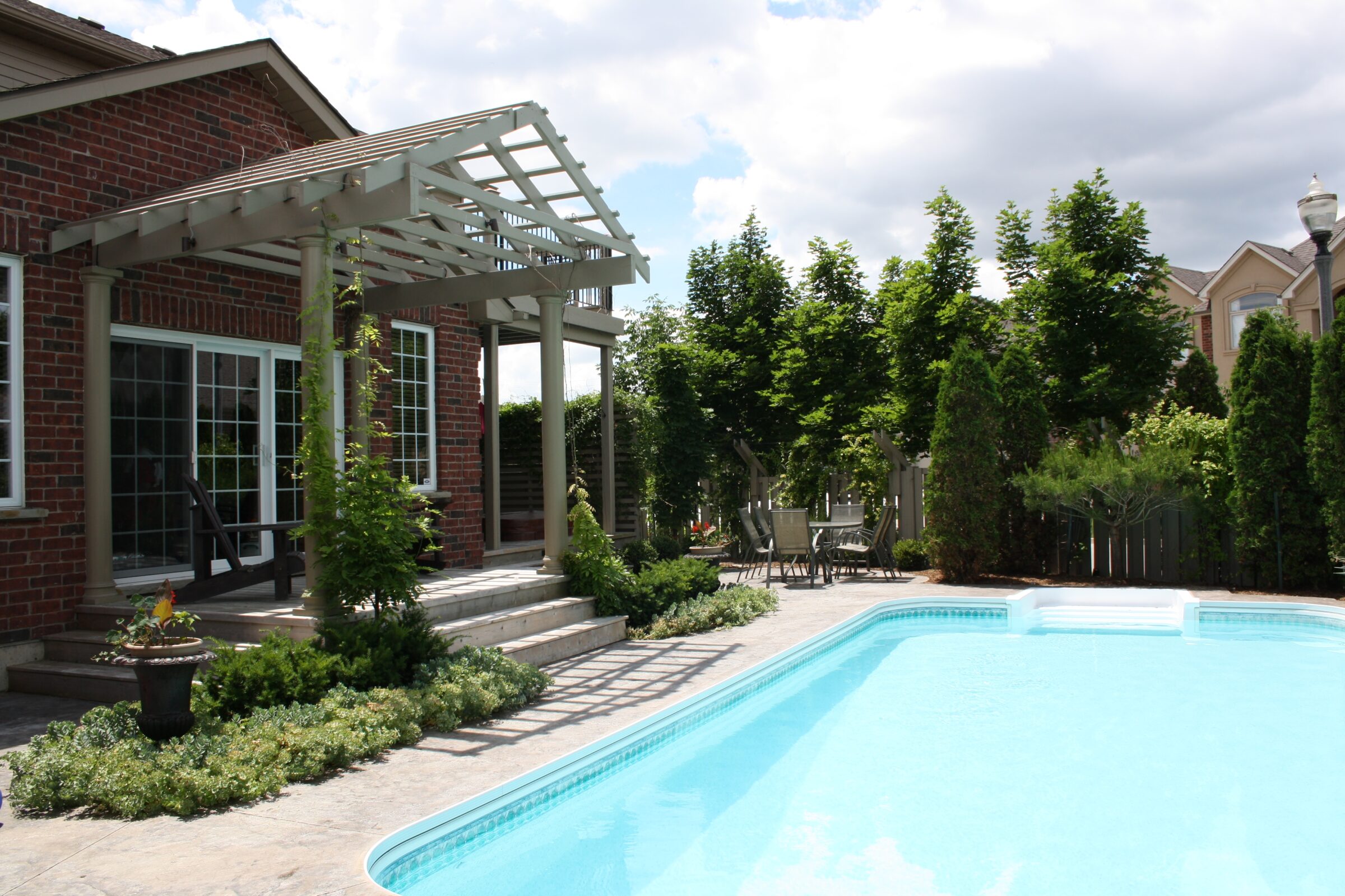 A backyard view of a red brick house with a pergola-covered patio, lush landscaping, and a sparkling in-ground swimming pool bordered by neatly trimmed greenery and tall privacy trees.