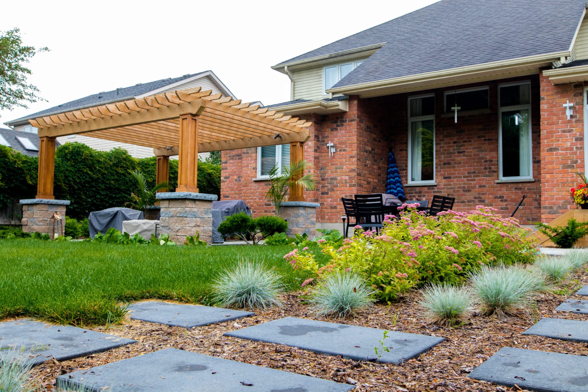 Backyard garden featuring pink spirea, ornamental switchgrass, and a modern pergola structure over a patio beside a red-brick home.