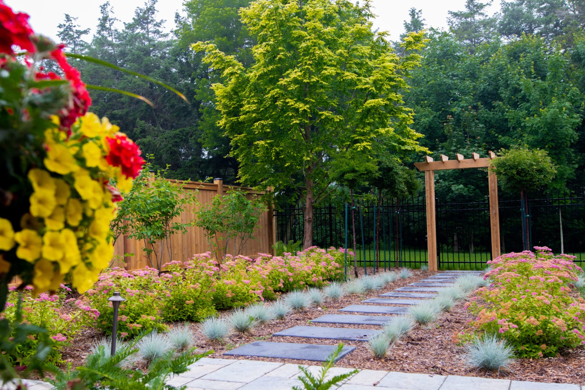Backyard garden with stone walkway, flowering plants, wooden arbor, and privacy fence.