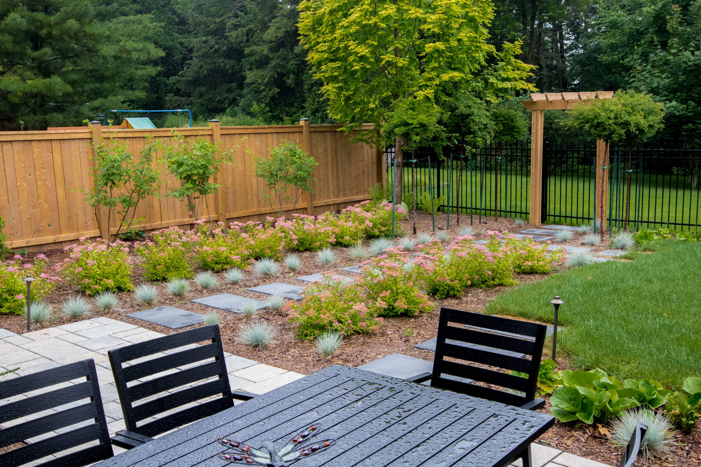 A backyard garden with structured planting beds, blooming pink flowers, ornamental grasses, and a stepping stone path leading to a wooden arbor and black metal fence. A black outdoor dining set sits in the foreground.