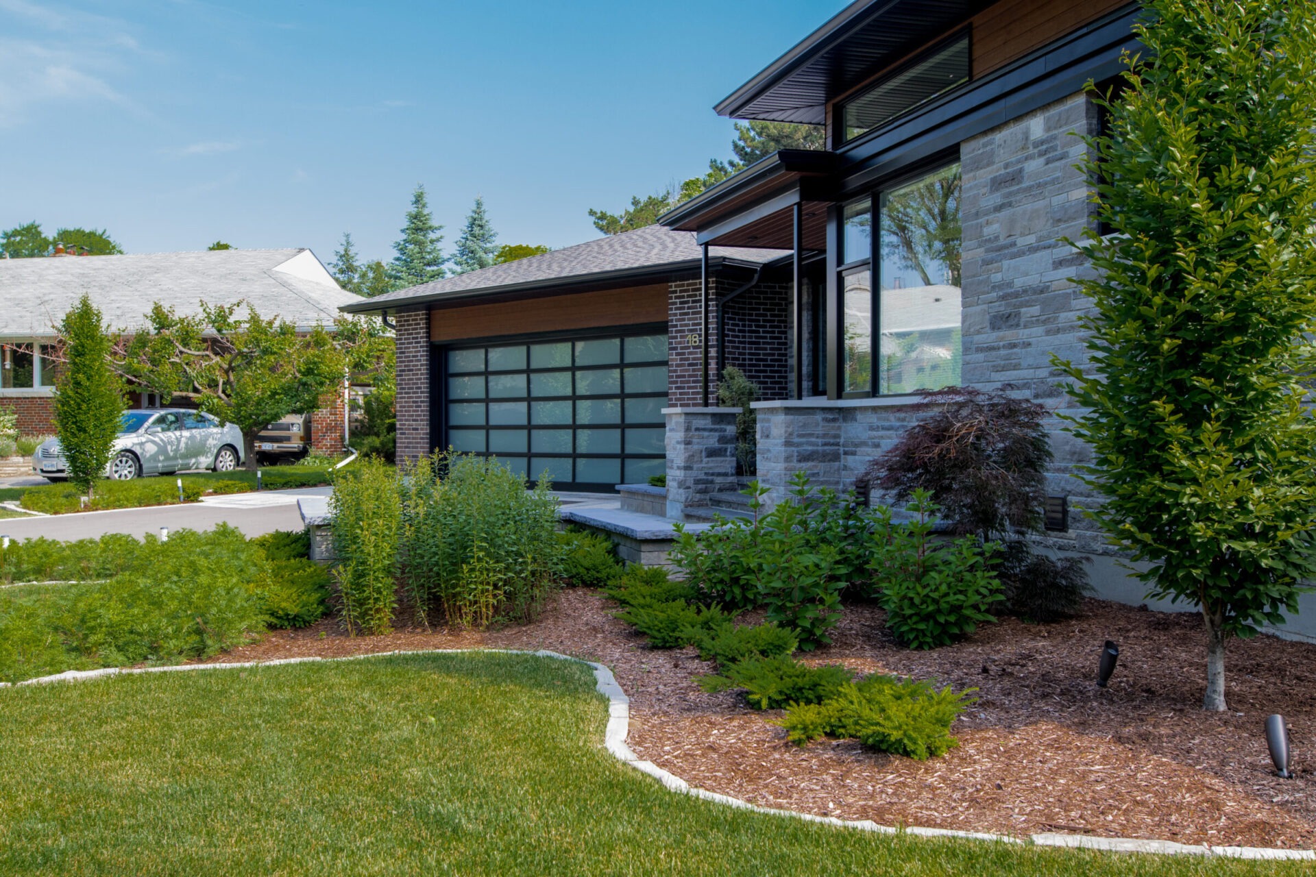 This image shows the front of a modern home with clean architectural lines and low-maintenance landscaping. The yard features well-manicured lawn edges, mulch beds with a mix of shrubs, ornamental grasses, and small trees. The overall design emphasizes a minimalist aesthetic with structured plant groupings and strong curb appeal.