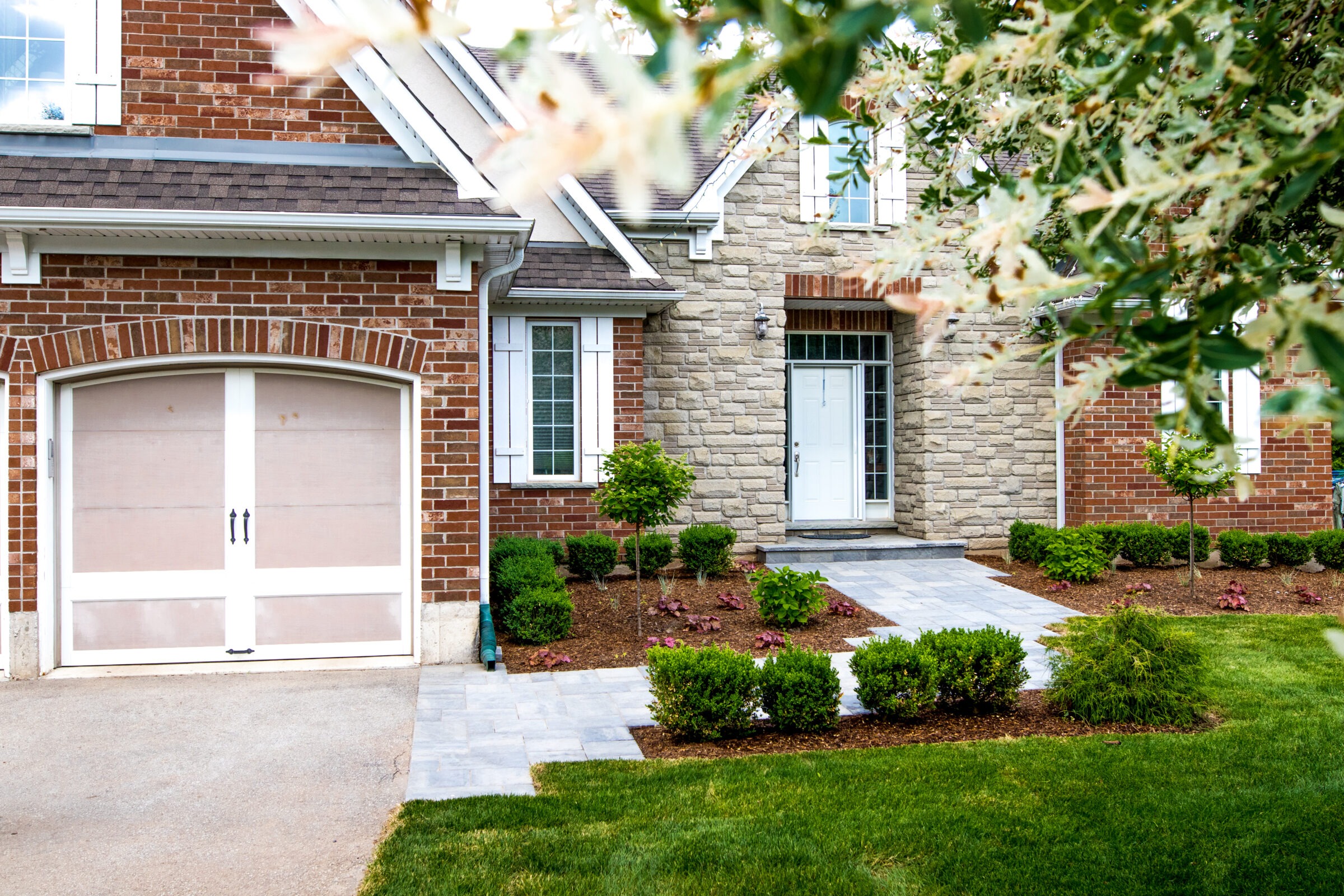 A well-maintained front yard with freshly edged garden beds, neatly trimmed shrubs, and a stone walkway leading to the front entrance of a brick and stone home with white trim and a white garage door. Blossoming tree branches frame the top of the image.