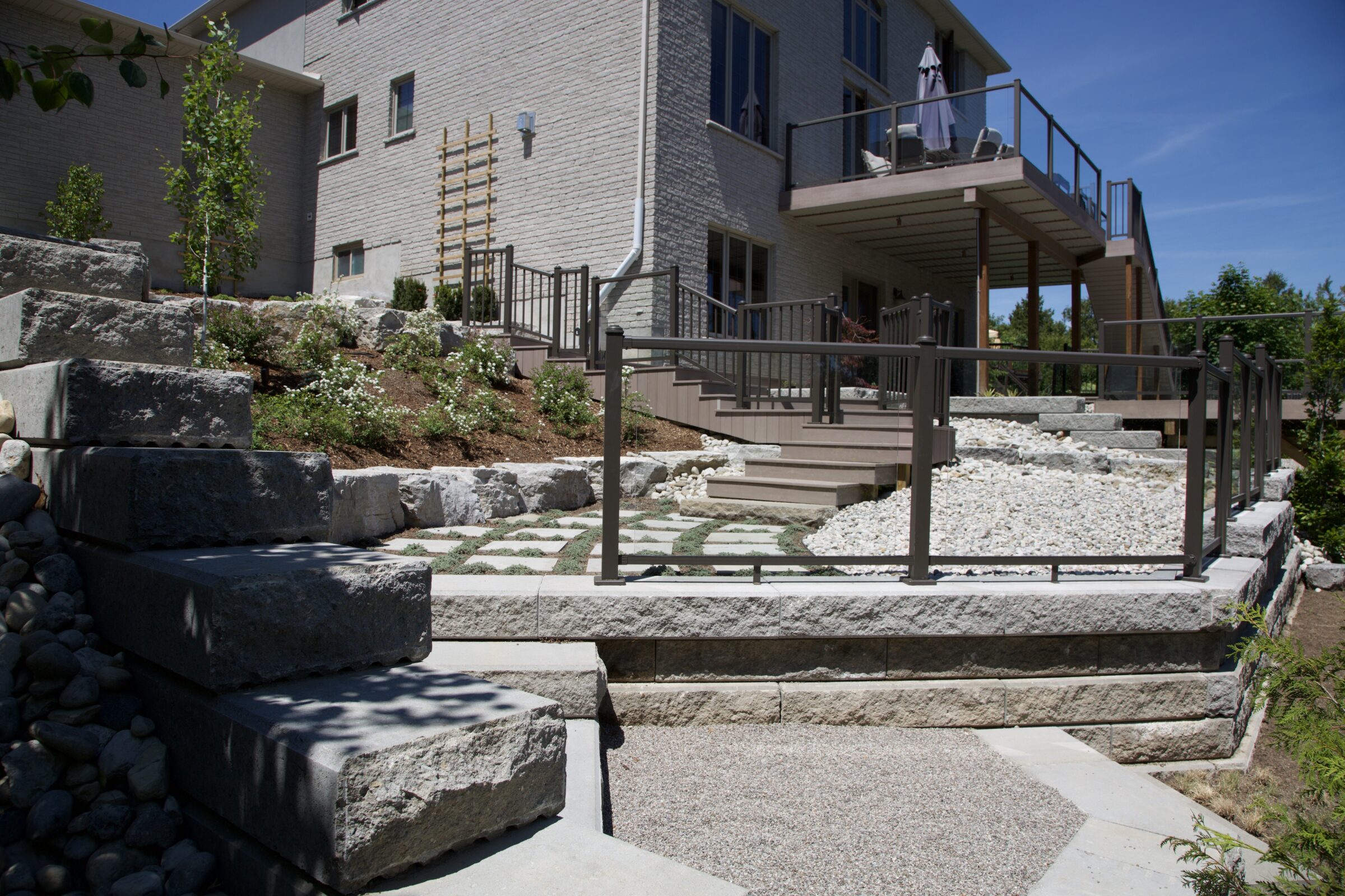 A contemporary sloped backyard design featuring layered retaining walls, stone slab steps, and a low-maintenance mix of groundcover, gravel, and ornamental plantings. Elevated decks with sleek railings provide multiple levels for outdoor entertaining and relaxation.