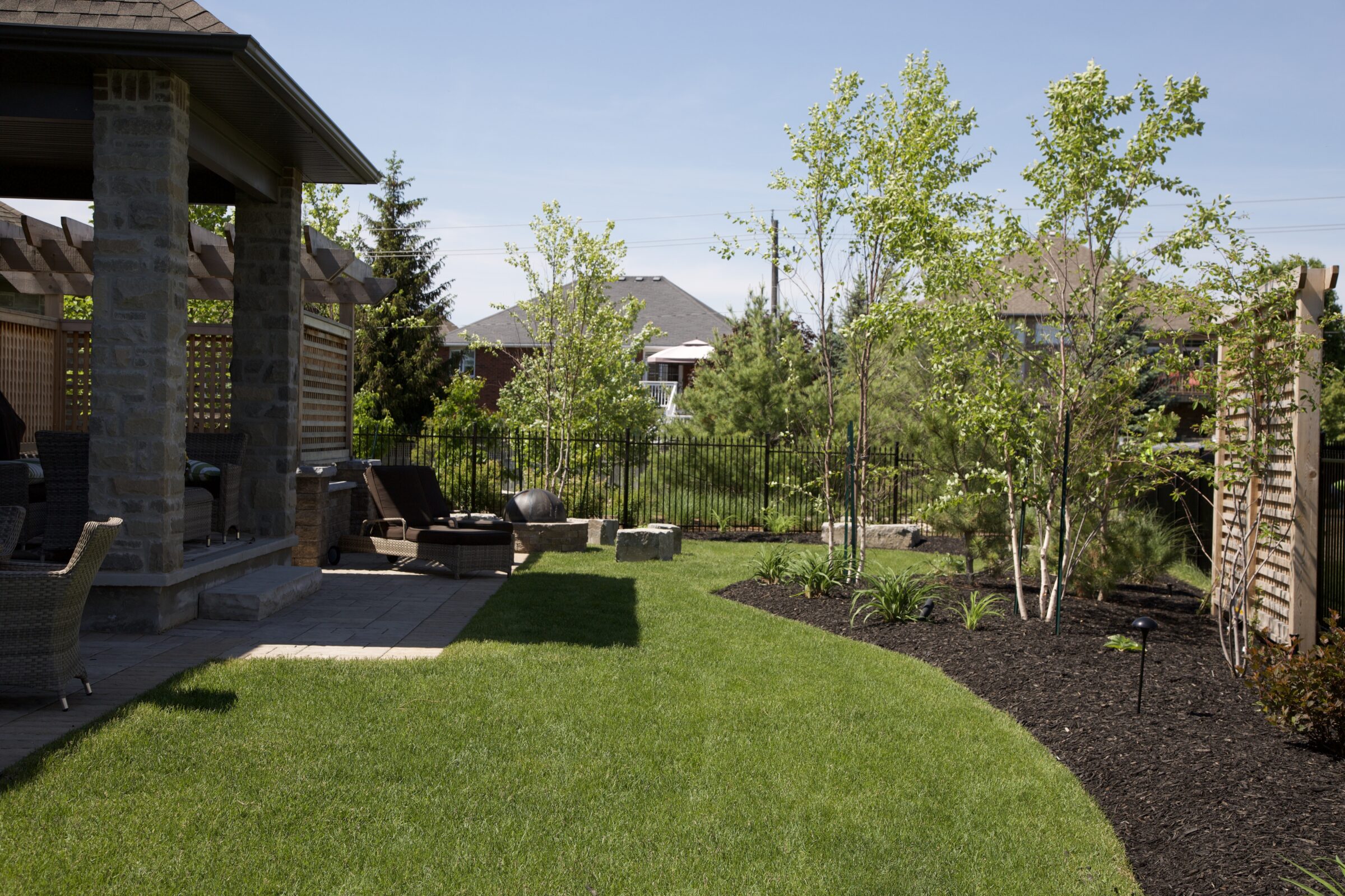 Backyard view featuring a covered stone patio with lounge seating, fresh green lawn, young trees along a mulched garden bed, and a wrought iron fence for privacy.