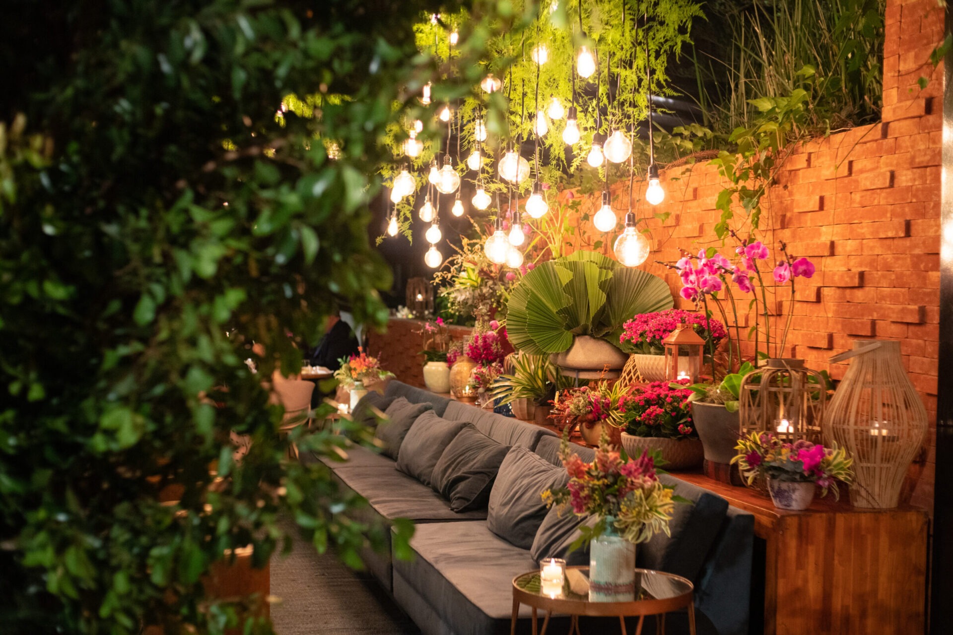 Outdoor patio lounge with hanging string lights, lush plants, and cozy seating against a brick wall.