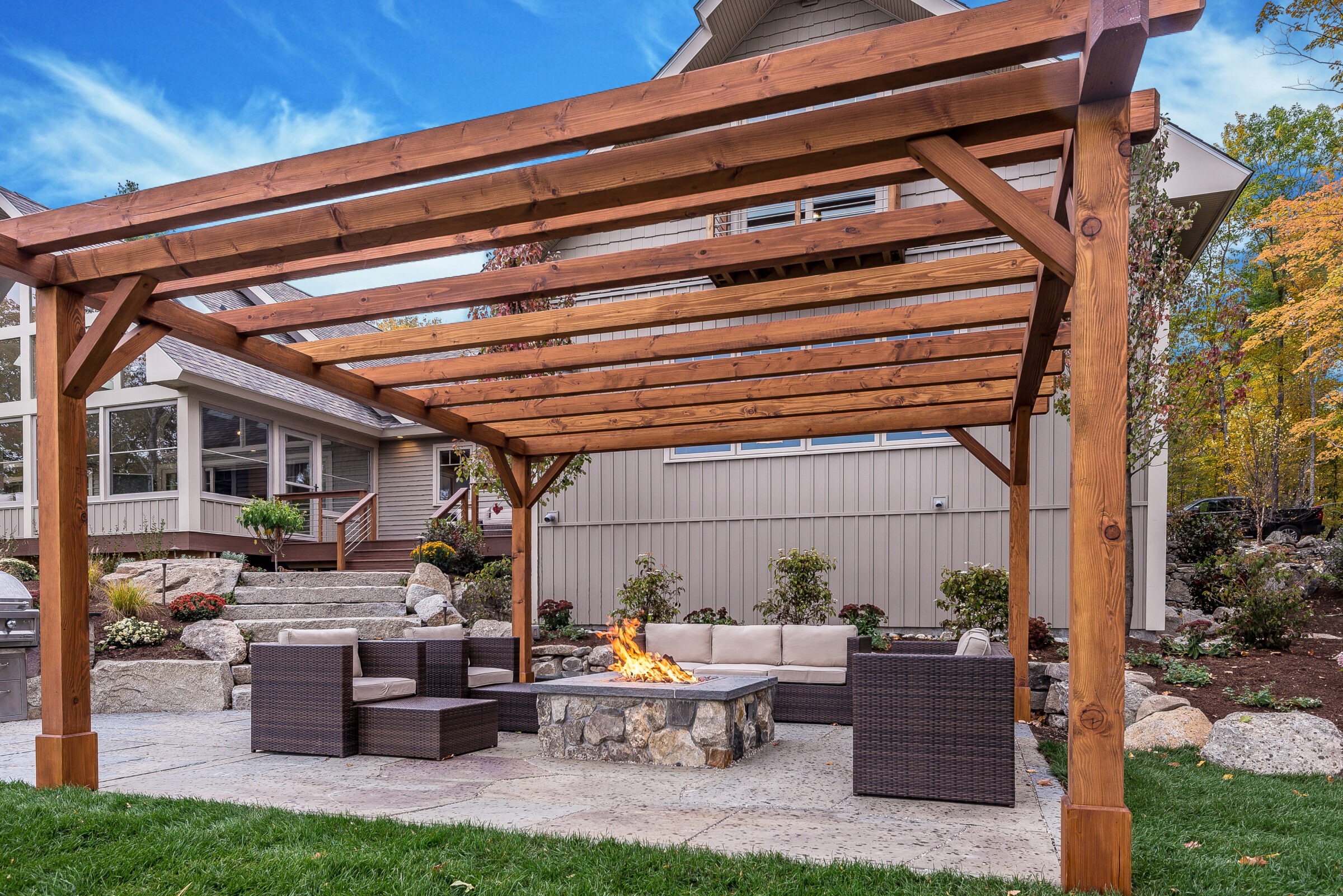 Backyard patio with a wooden pergola, stone fire pit, and wicker outdoor seating surrounded by landscaping.