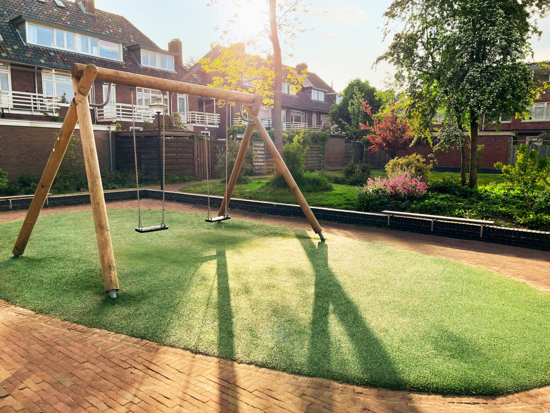 A small community playground with a minimalist natural wood swing set on a soft turf surface, surrounded by brick paving and lush garden beds. The golden hour sunlight and blooming shrubs give the area a peaceful, family-friendly vibe.