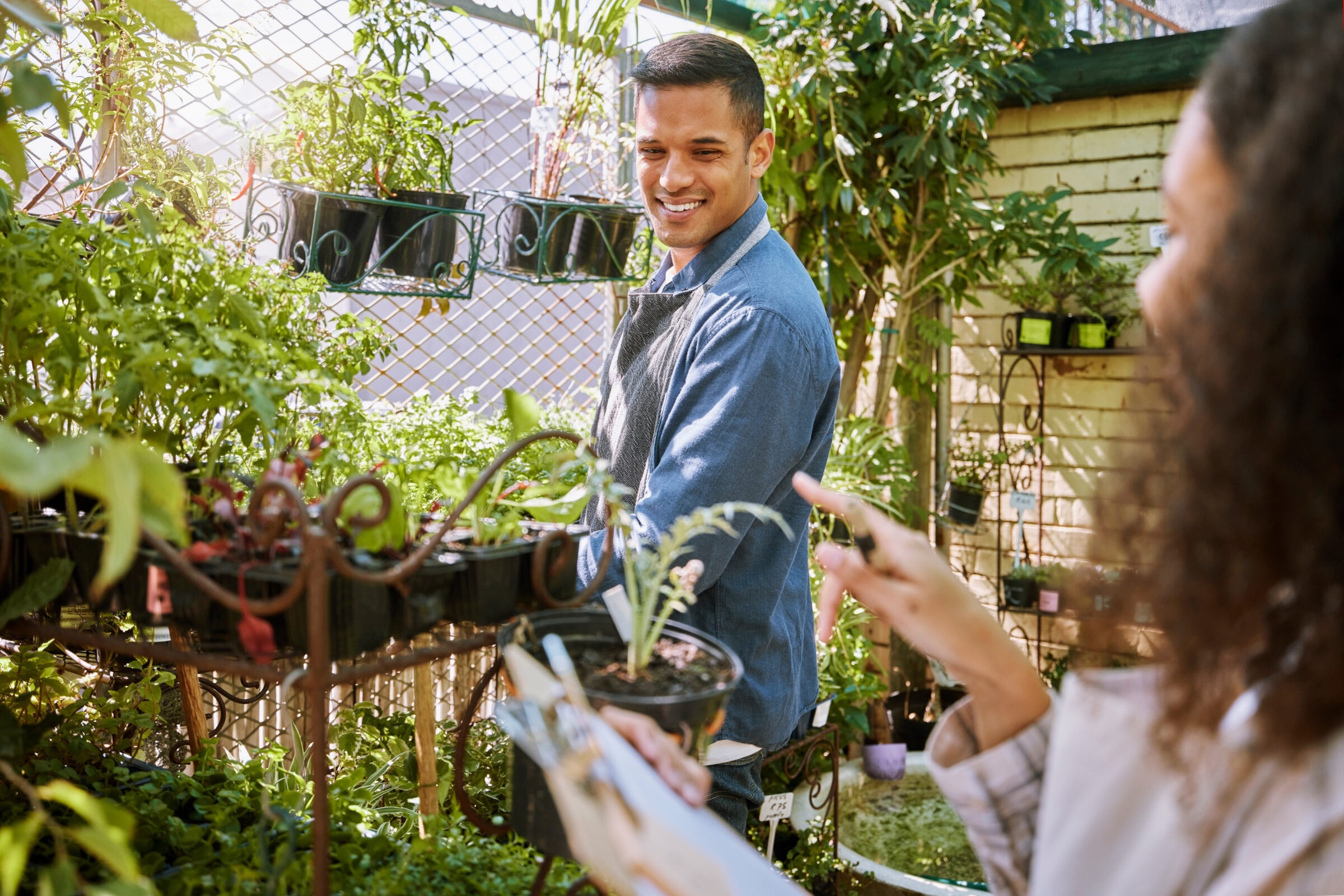Two people interacting in a lush garden center or nursery. A man in a denim shirt and apron smiles while tending to potted plants, as a woman in the foreground points toward a plant while holding a clipboard, possibly discussing a gardening plan or plant selection.