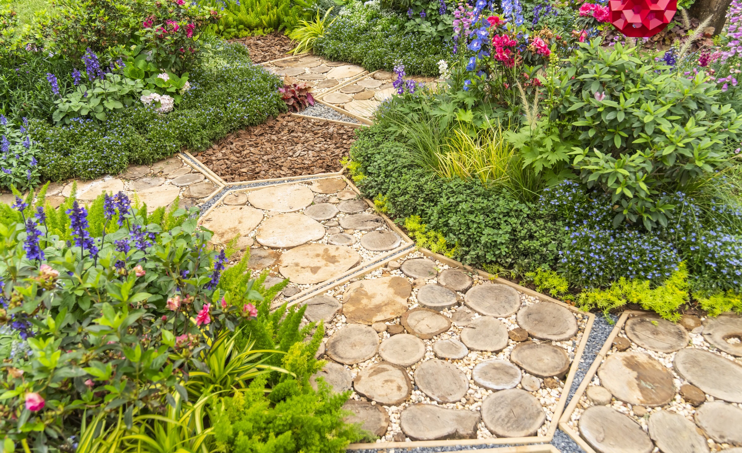 A whimsical garden pathway made of circular wood slice pavers set in gravel, weaving through lush, colorful flower beds filled with vibrant blooms, greenery, and groundcovers.