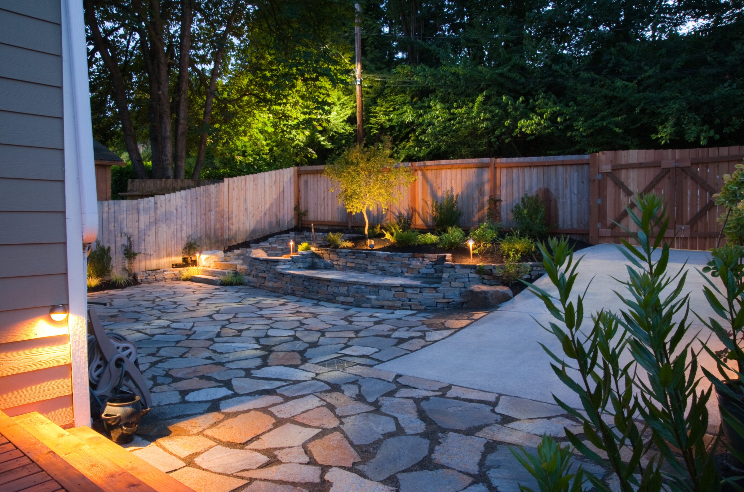 Evening backyard patio with flagstone paving, tiered stone retaining walls, soft landscape lighting, and a central tree as a focal point against a wood privacy fence.
