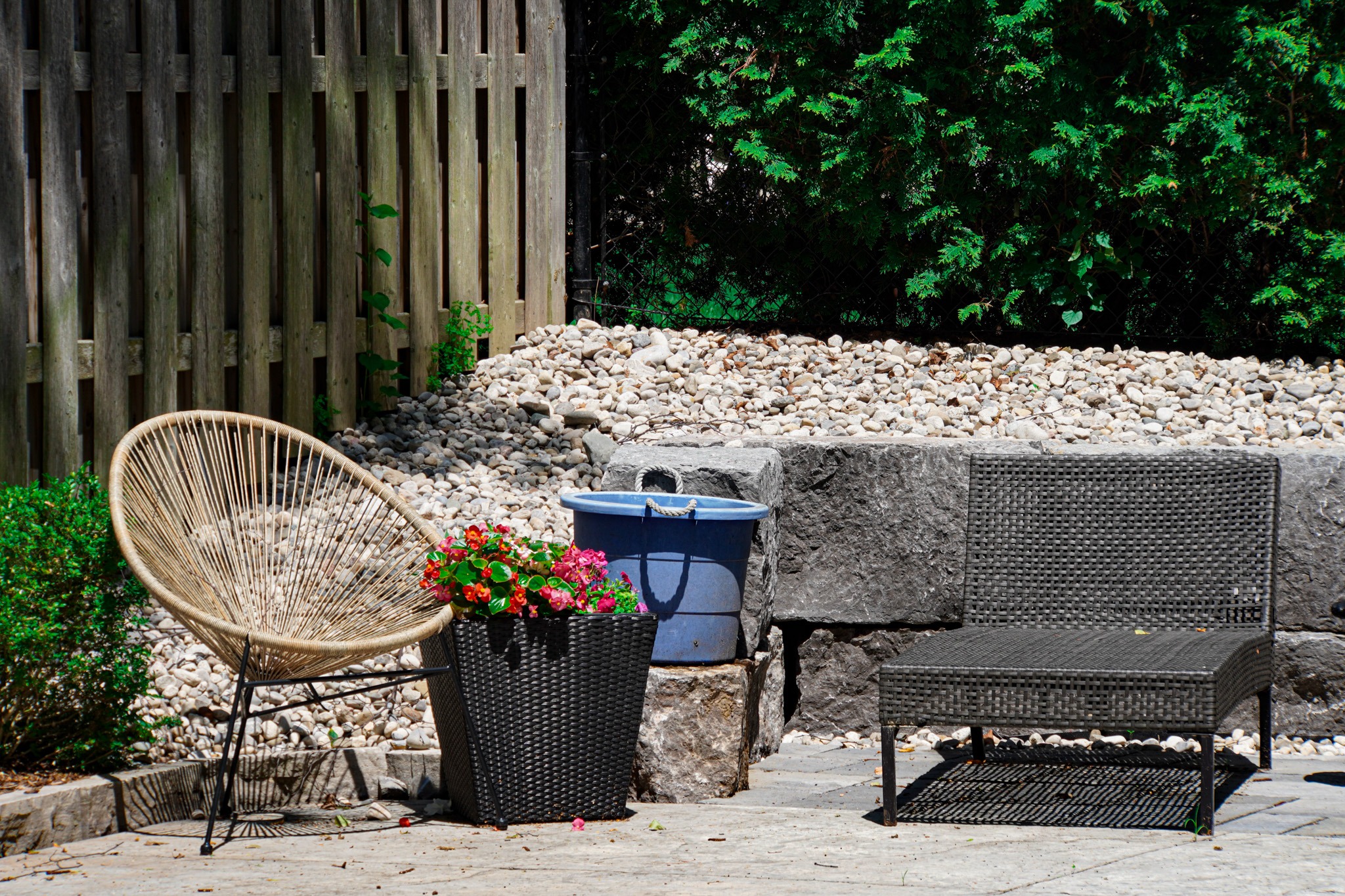 Cozy backyard corner with two modern outdoor chairs—one round woven and the other square wicker—next to a tall planter filled with red and pink flowers, a blue bucket, and a backdrop of stone blocks, river rocks, a wooden fence, and evergreen trees.