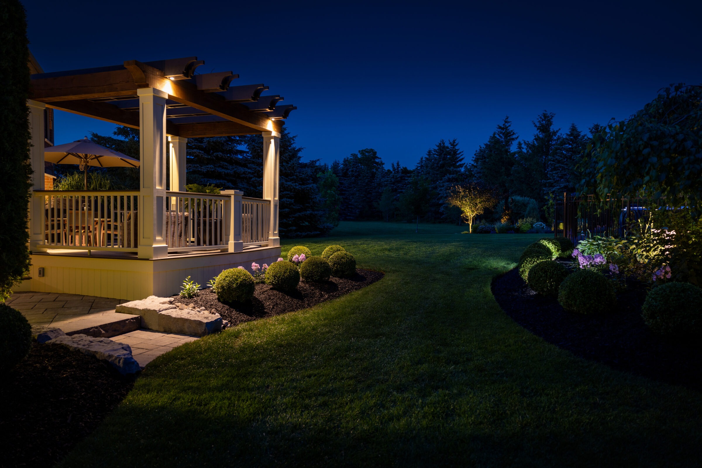 Illuminated backyard patio with a wooden pergola and cozy seating, surrounded by manicured shrubs and garden beds. Landscape lighting highlights trees and flower beds across a spacious lawn under a dark evening sky.