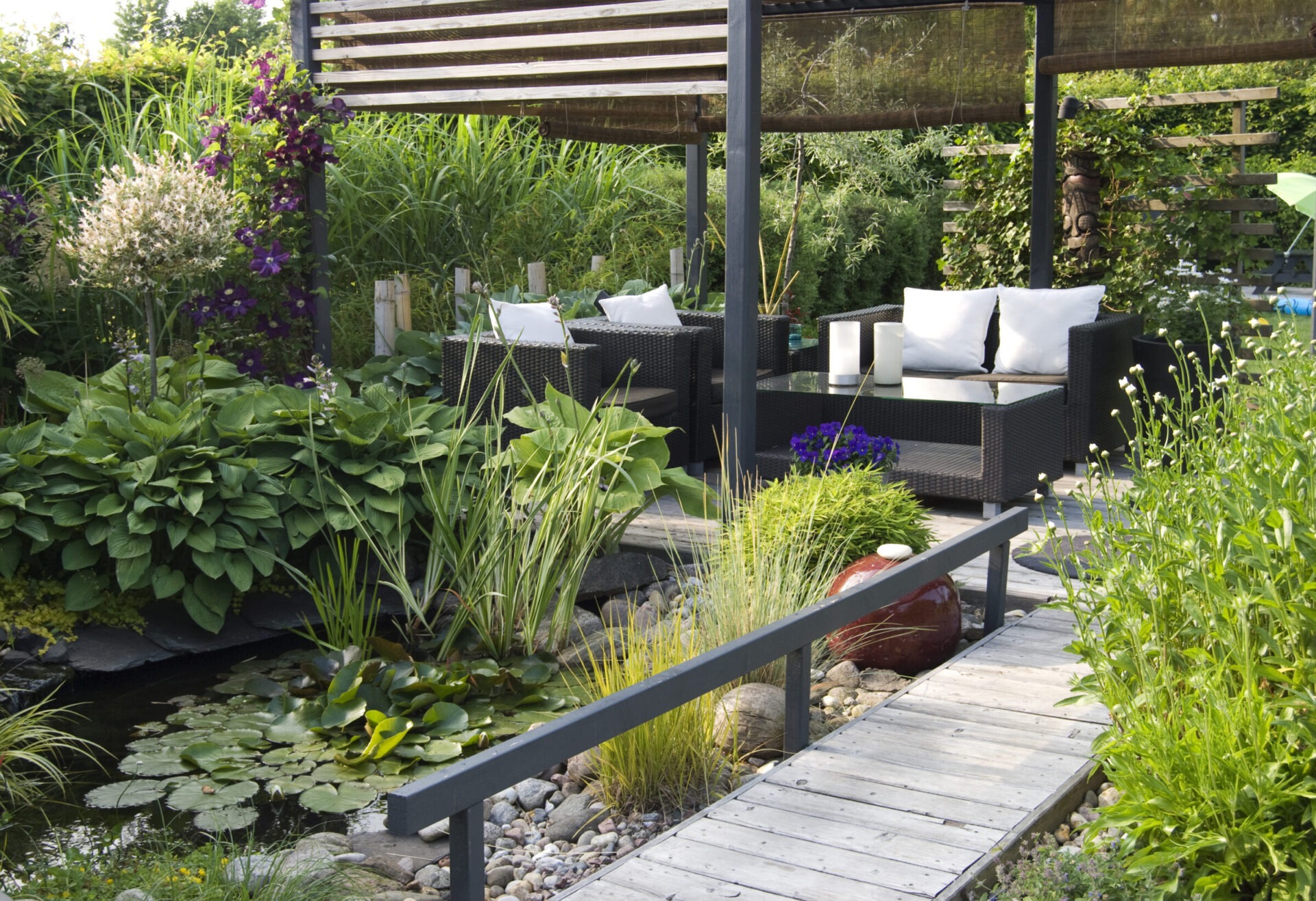 Serene backyard patio with a modern pergola, wicker seating, and a glass coffee table, overlooking a pond filled with lily pads and surrounded by lush plants. A wooden walkway with railing leads through the garden.
