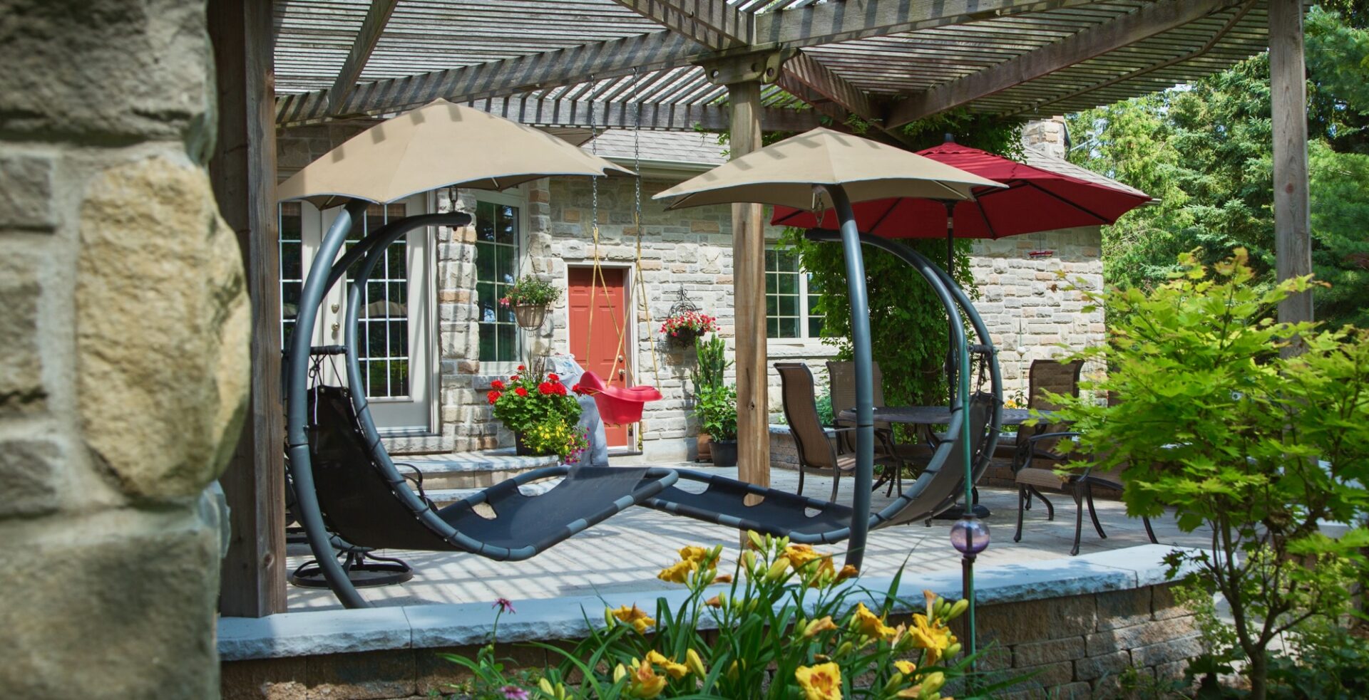 Stone patio featuring a wooden pergola, circular loungers with umbrellas, a red child’s swing, and colorful potted flowers. Surrounded by greenery and set against a brick home exterior.