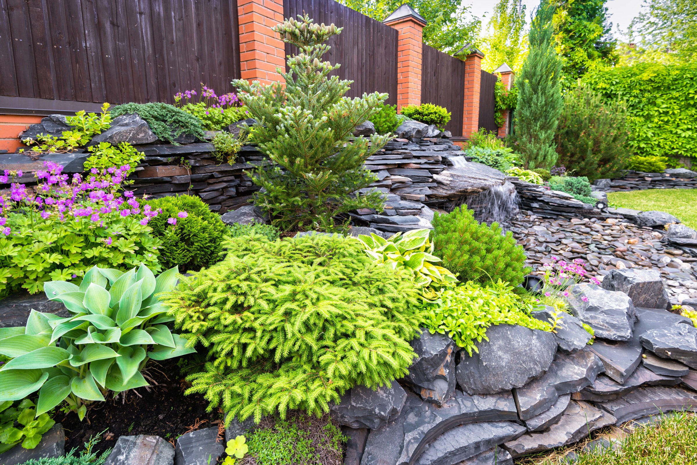 Beautifully landscaped rock garden with layered slate stones, evergreen shrubs, hostas, and a small cascading waterfall. Surrounded by colorful flowers and enclosed by a dark wooden fence with brick columns.