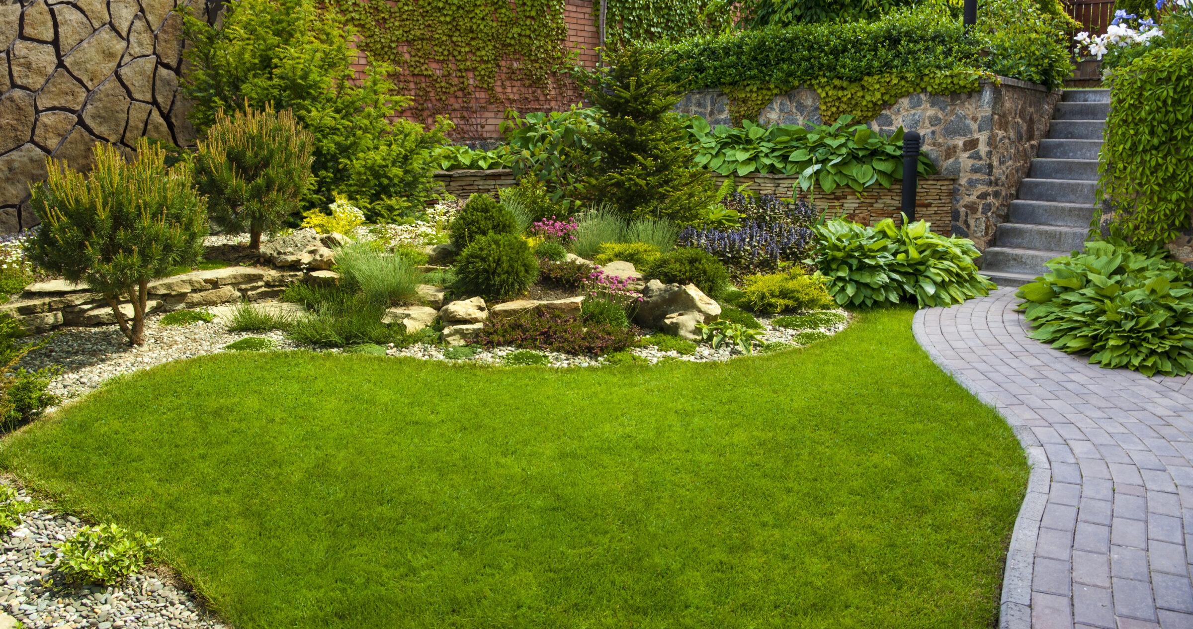 Beautifully landscaped garden featuring a curved brick walkway, manicured green lawn, layered stone borders, and a mix of evergreens, flowering perennials, and hostas, with ivy-covered walls and stone steps leading to an elevated area.
