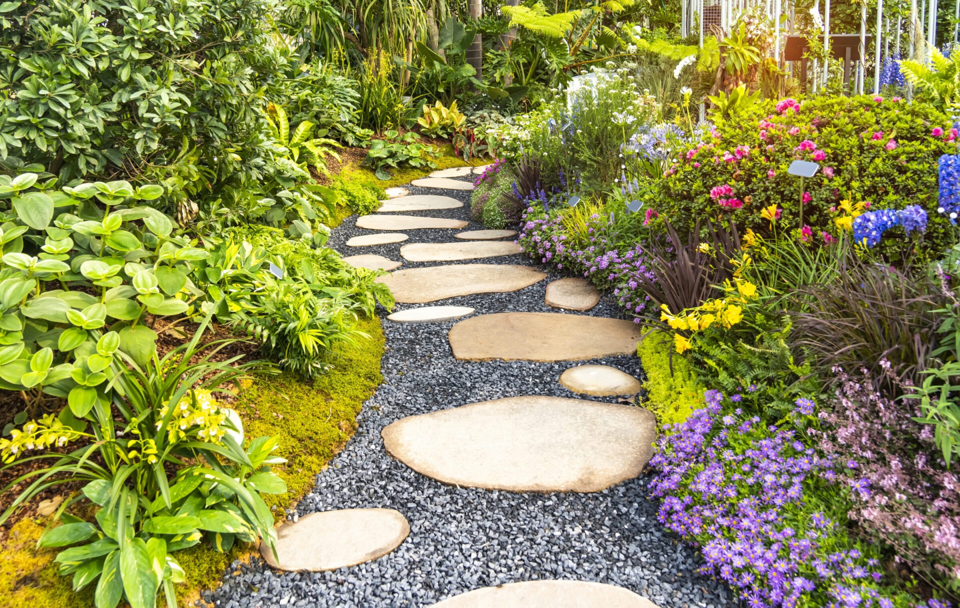 Vibrant garden pathway with large stepping stones set in dark gravel, winding through lush greenery and blooming flowers in shades of pink, purple, yellow, and blue, creating a colorful and inviting landscape scene.