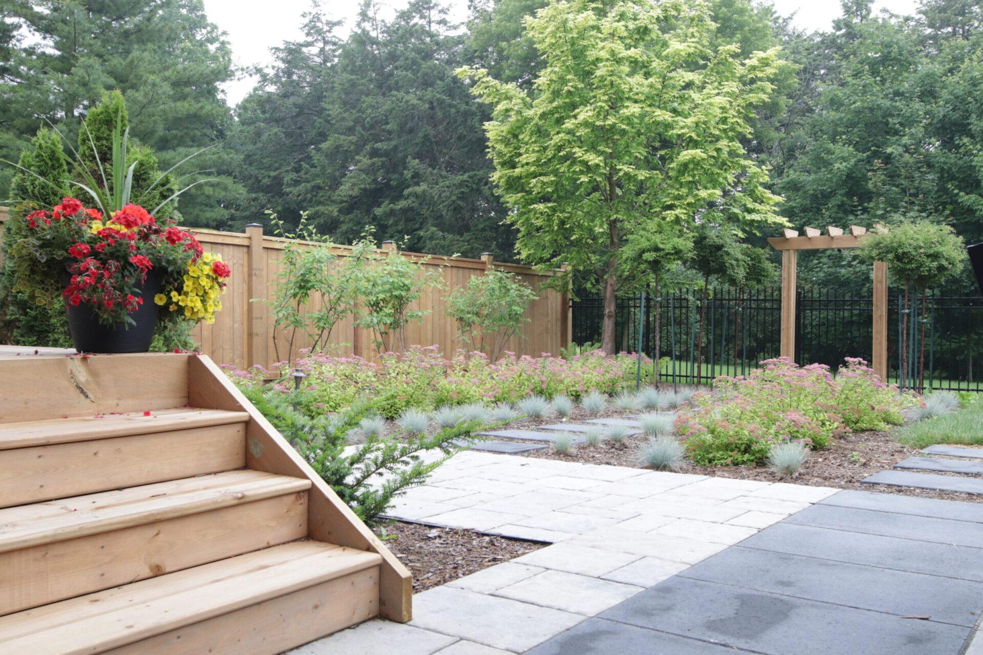 Backyard garden with wooden steps leading to a patio, bordered by potted flowers, stone pathways, ornamental grasses, blooming shrubs, and a wooden arbor beside a fenced tree-lined y
