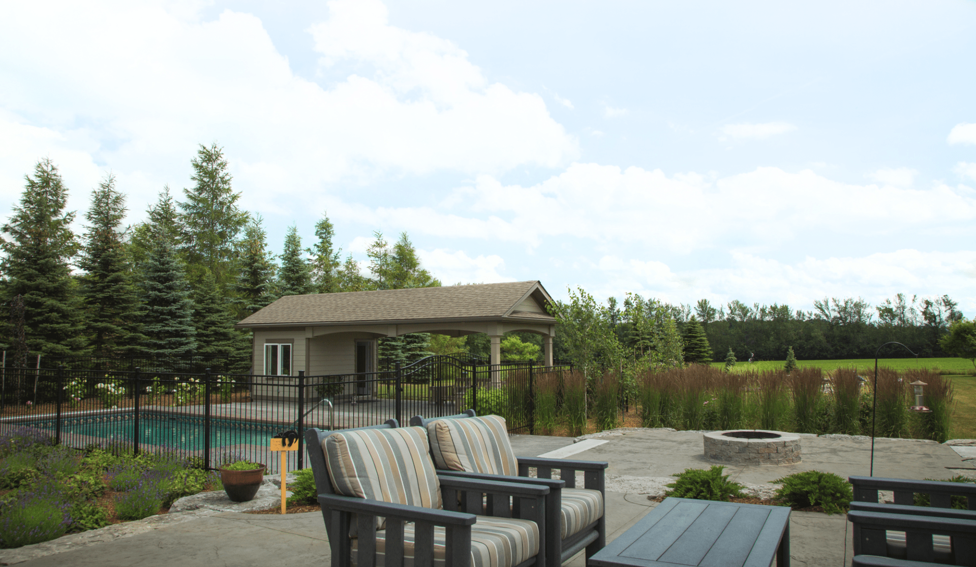 Backyard patio with cushioned chairs near a fire pit, overlooking a fenced swimming pool and pool house surrounded by tall evergreens.