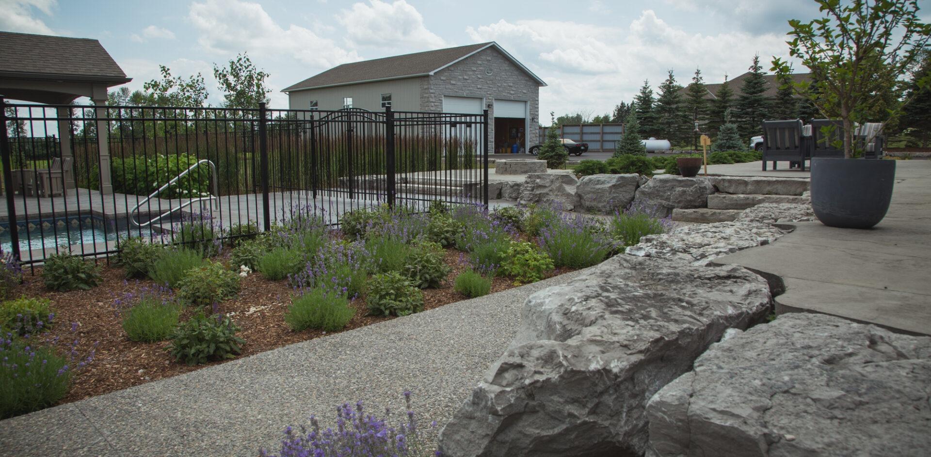 Landscaped backyard with a fenced pool area, lavender garden beds, large natural stone outcrops, and a seating patio near a stone outbuilding.