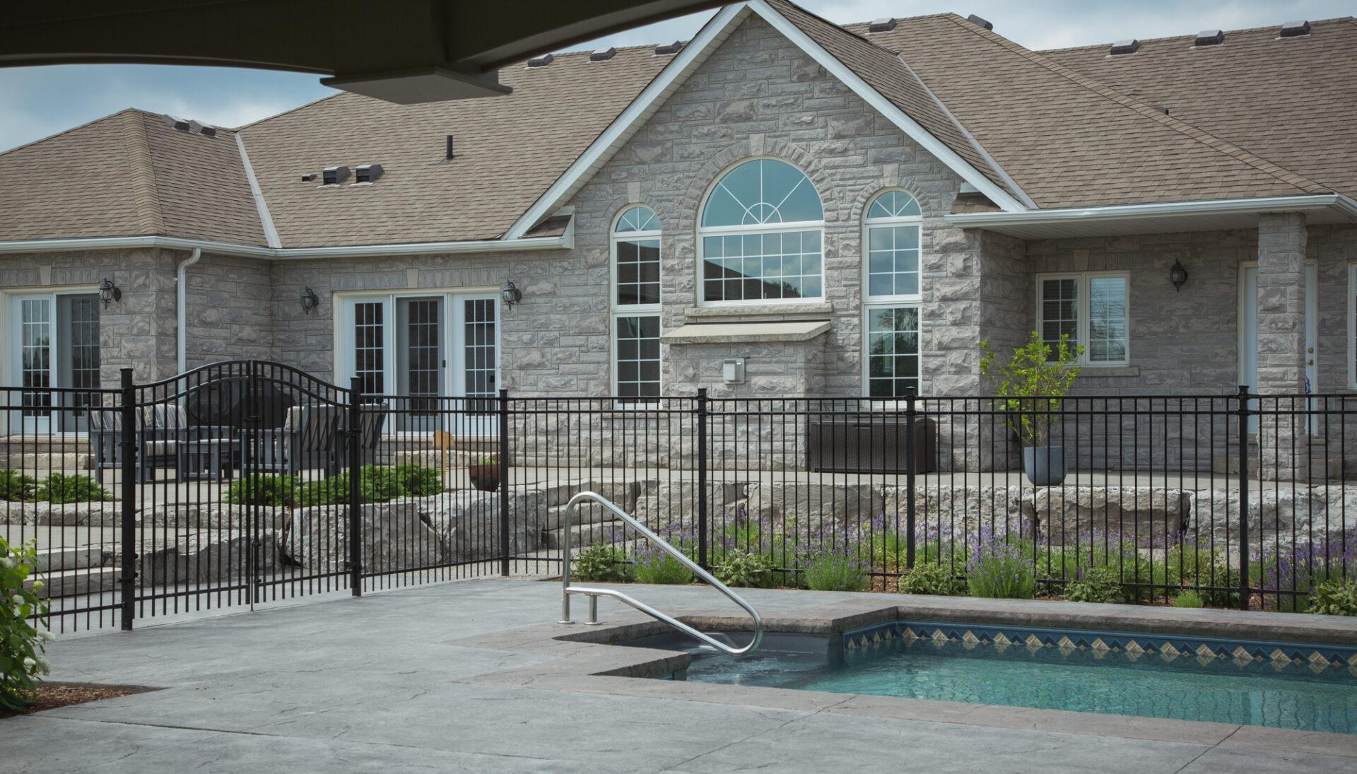 Pool area with black railing, stone patio, and landscaping beds in front of a large stone house with multiple windows and doors.