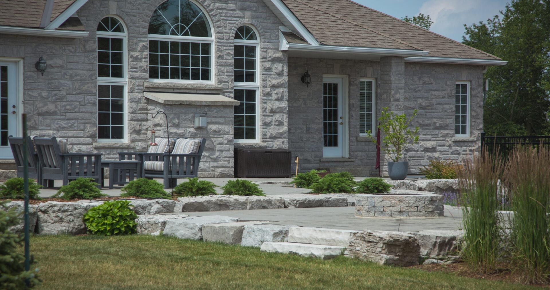 Stone patio with seating and a circular fire pit bordered by layered garden beds and natural stone steps leading to a large home.