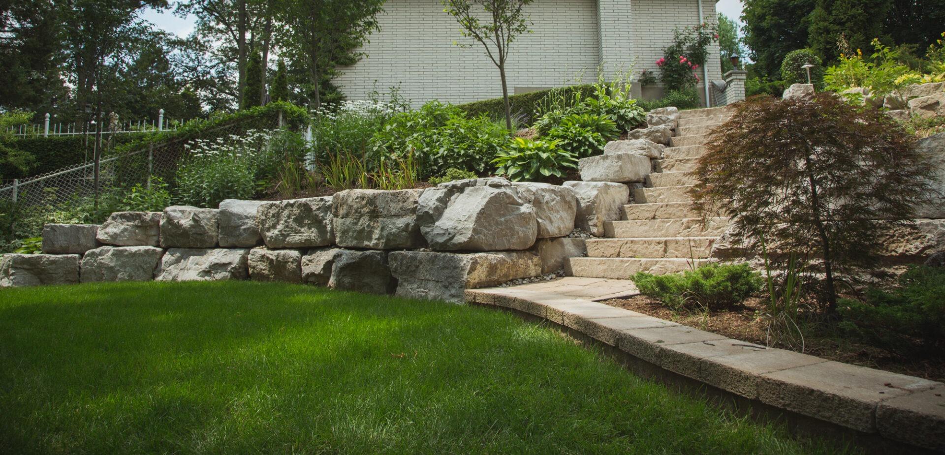 Tiered backyard with large stone retaining walls, lush plantings, and wide natural stone steps leading to the upper garden near a white brick home.